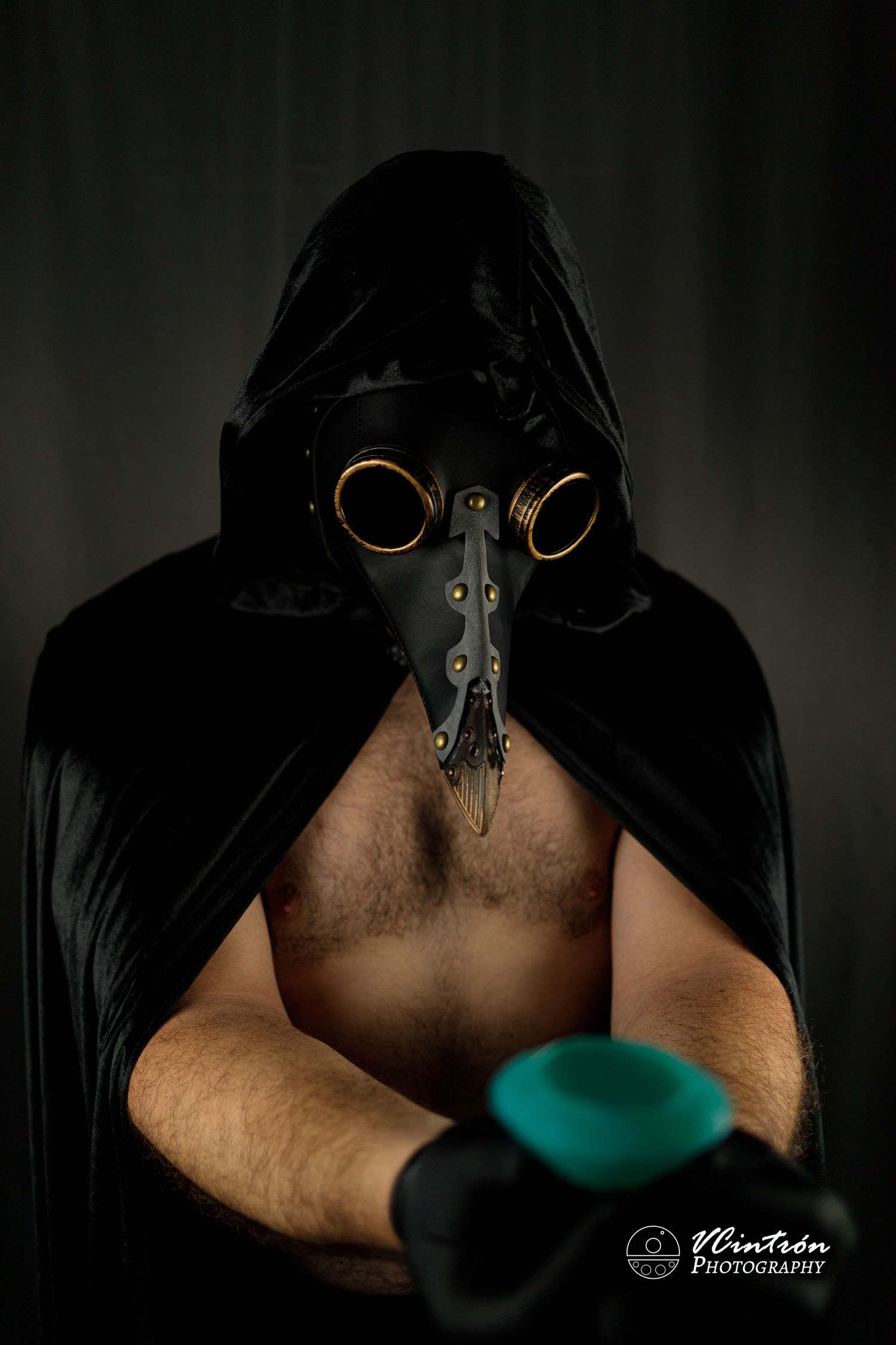 Dark and moody portrait of a plague doctor wearing a black hooded cloak and steampunk leather mask with bronze goggles.