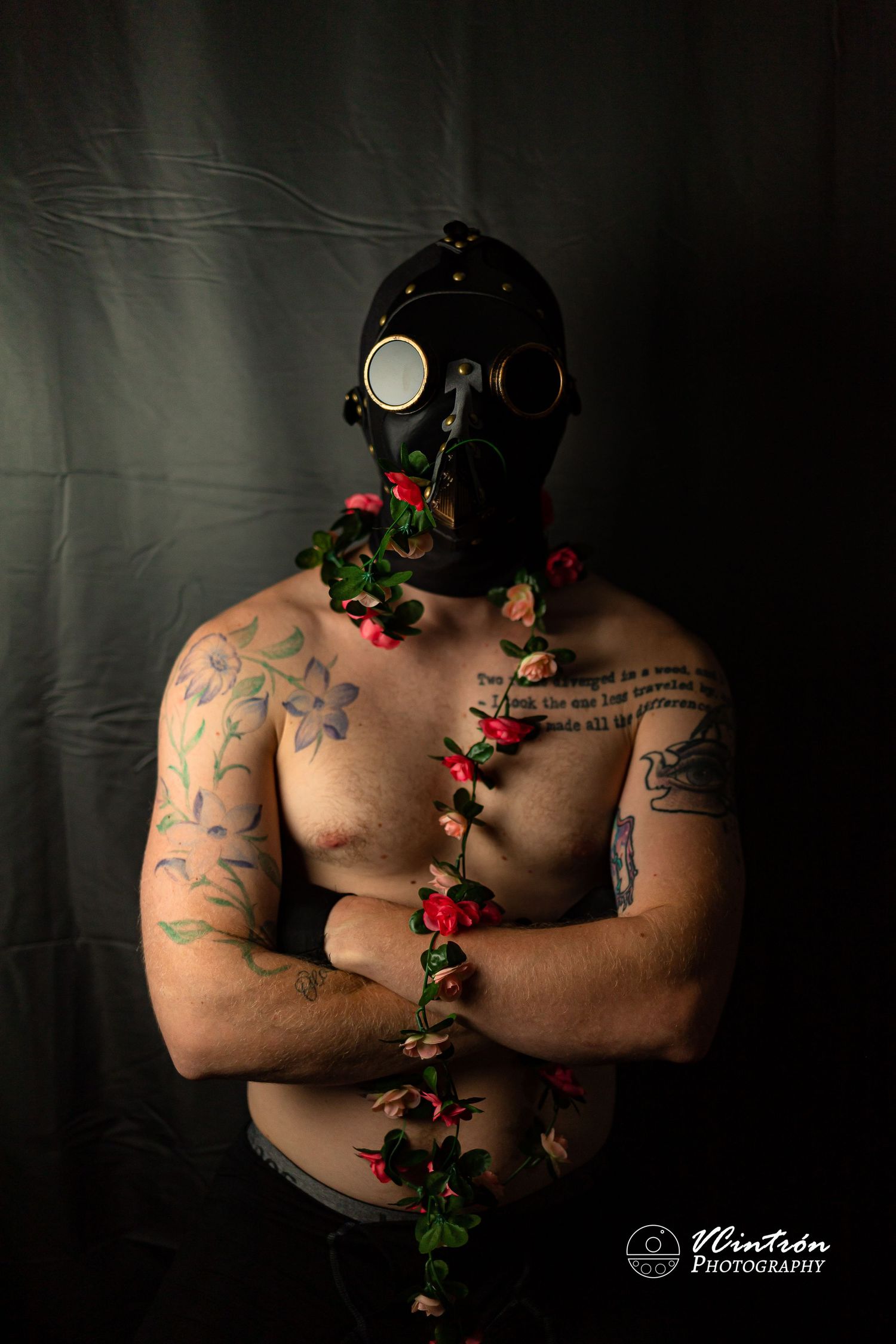 Artistic portrait of shirtless tattooed figure wearing black gas mask against dark backdrop with red roses and vines.