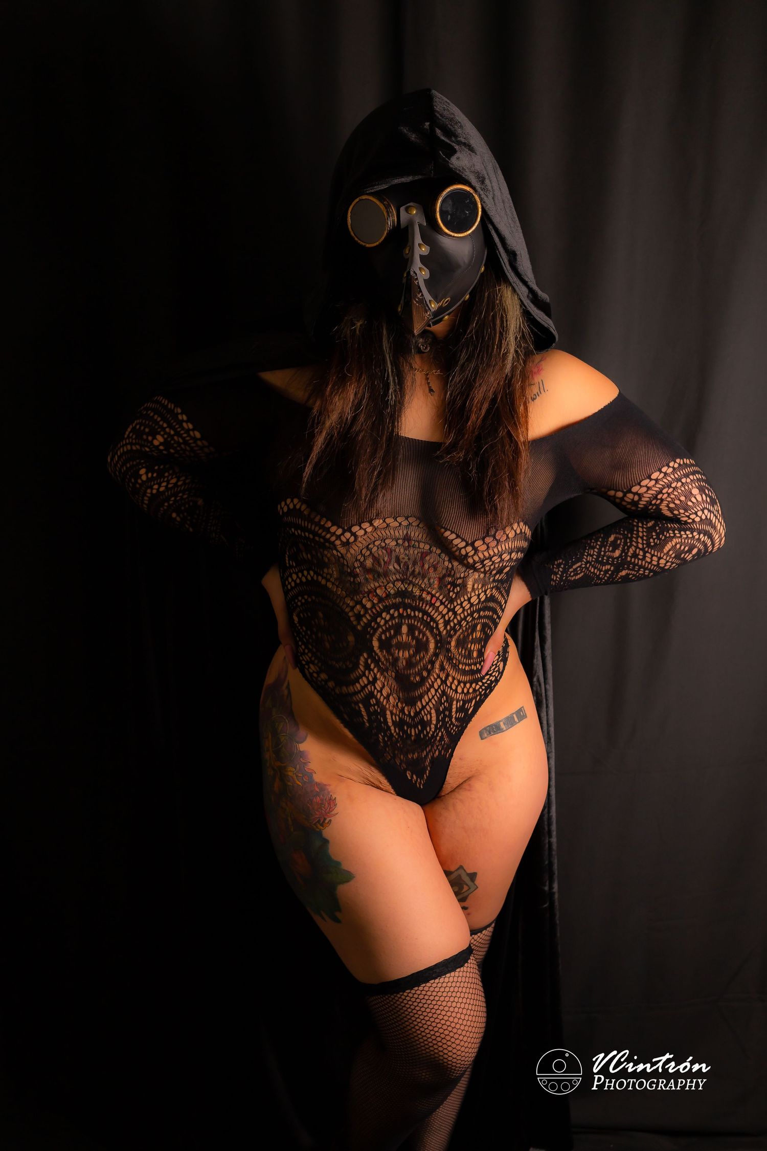 Dark artistic portrait of a figure in black lace bodysuit, gas mask and steampunk goggles against black backdrop.