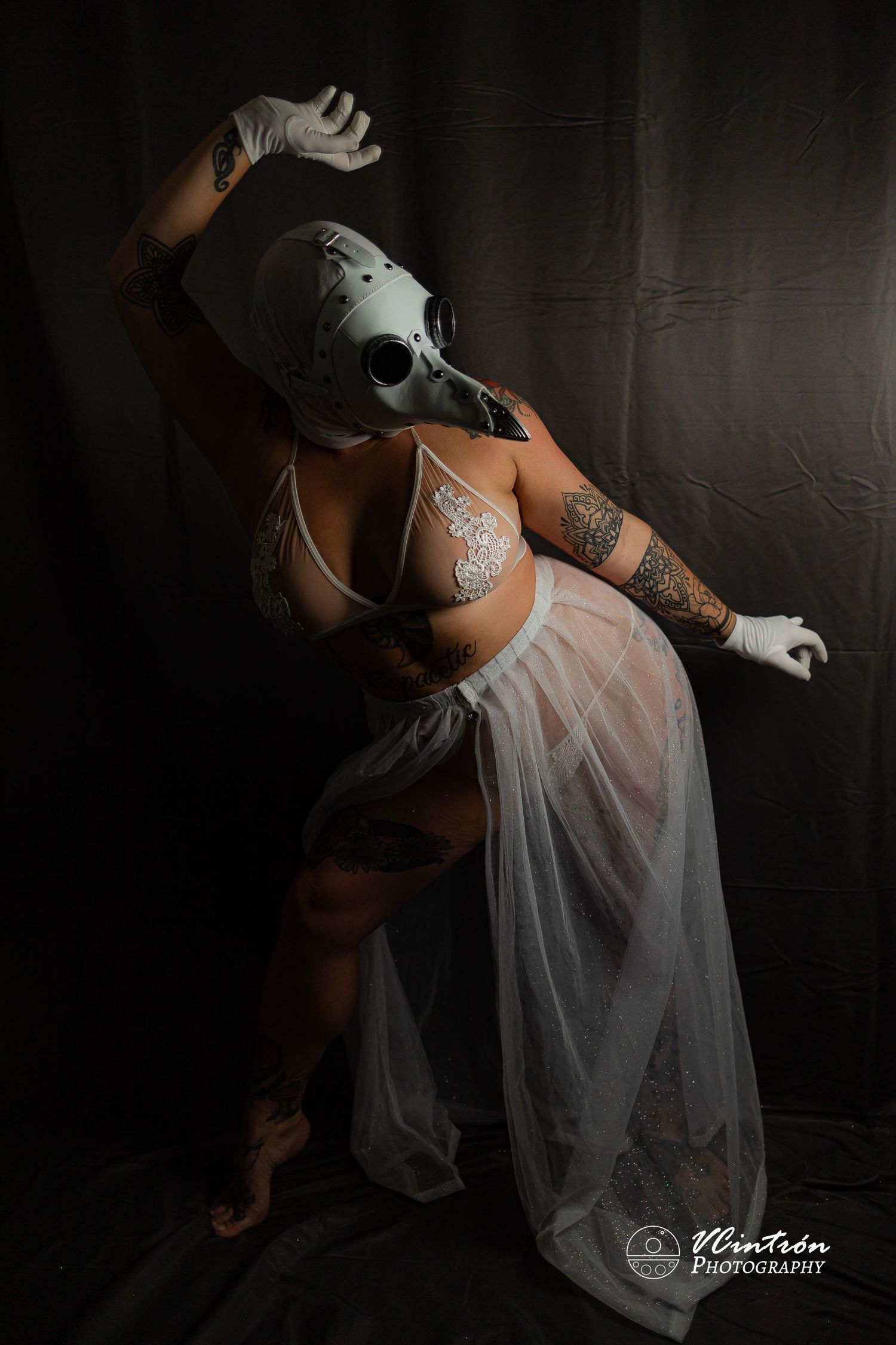 A dancer in white lingerie and plague doctor mask performs an artistic pose against a dark backdrop.