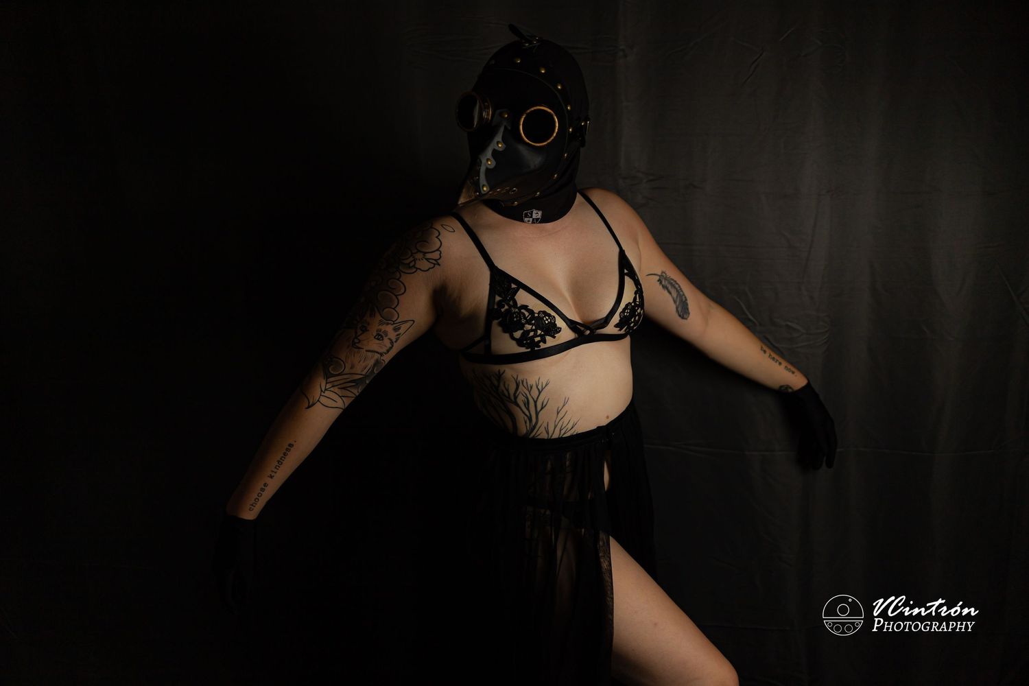 Figure in black gas mask and lingerie poses against dark background in moody artistic portrait.