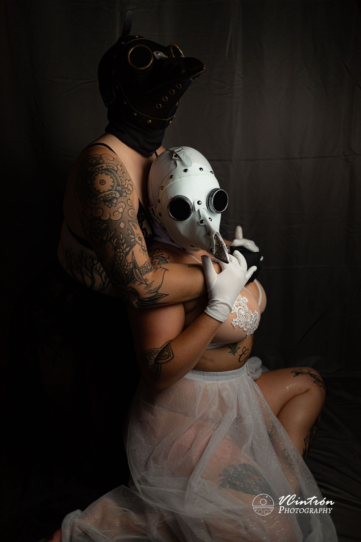 Dark atmospheric portrait with tattooed figure in black gas mask and another in white hockey mask with sheer dress in moody lighting.