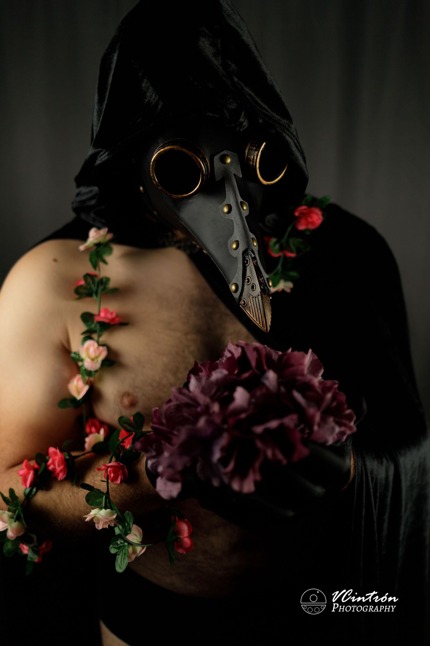 Dark and moody portrait featuring a plague doctor mask, black cloak, and purple flowers with delicate red blooms against shadows.