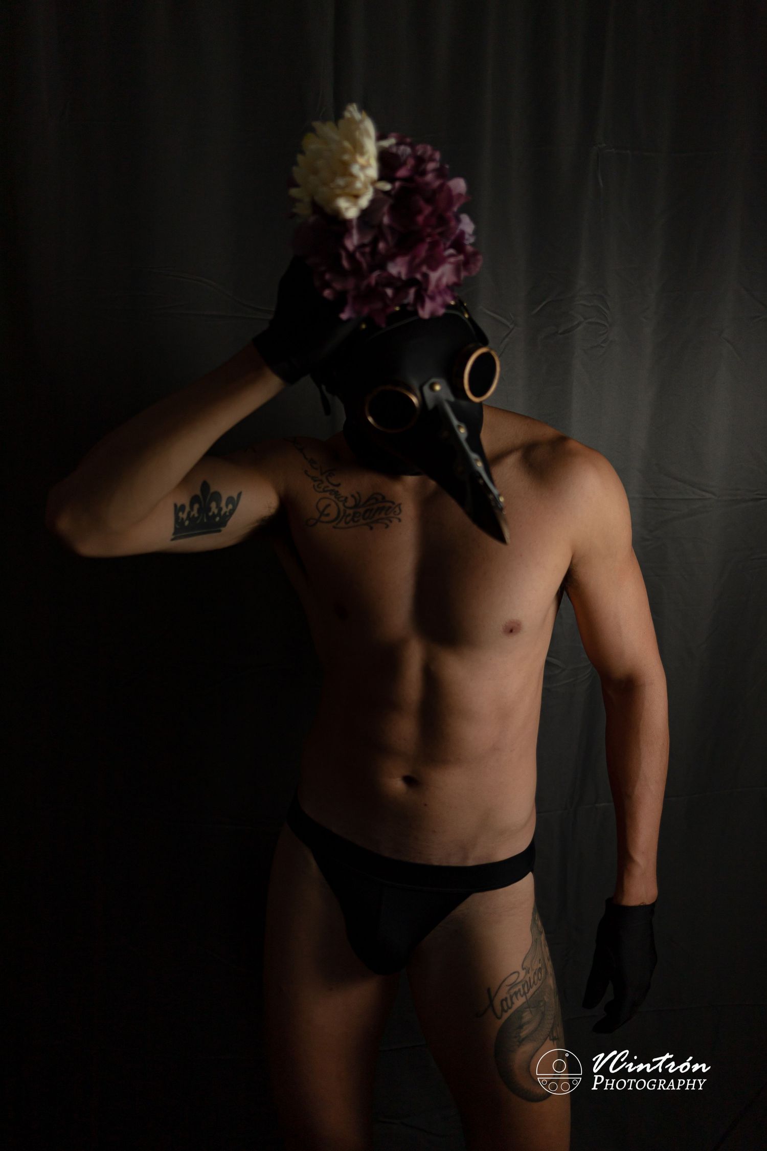 Artistic boudoir photo with black plague doctor mask adorned with purple and cream flowers in moody lighting.