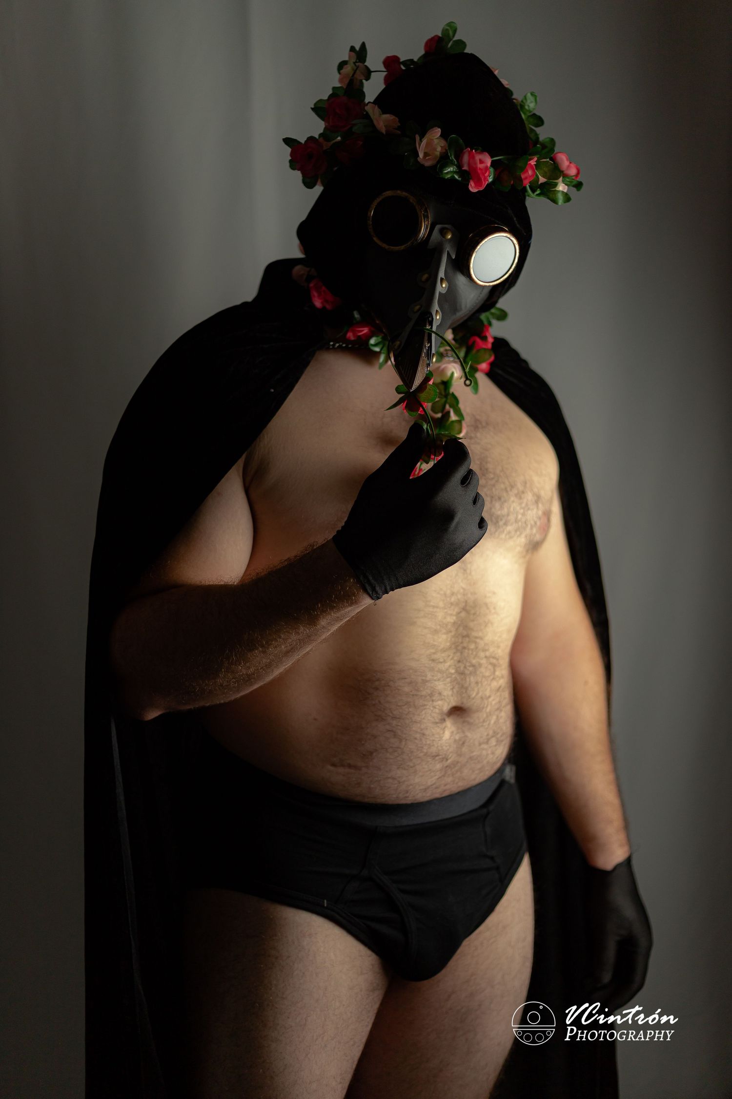 Artistic portrait of person in black gas mask adorned with red flowers and dark outfit against shadowy backdrop.