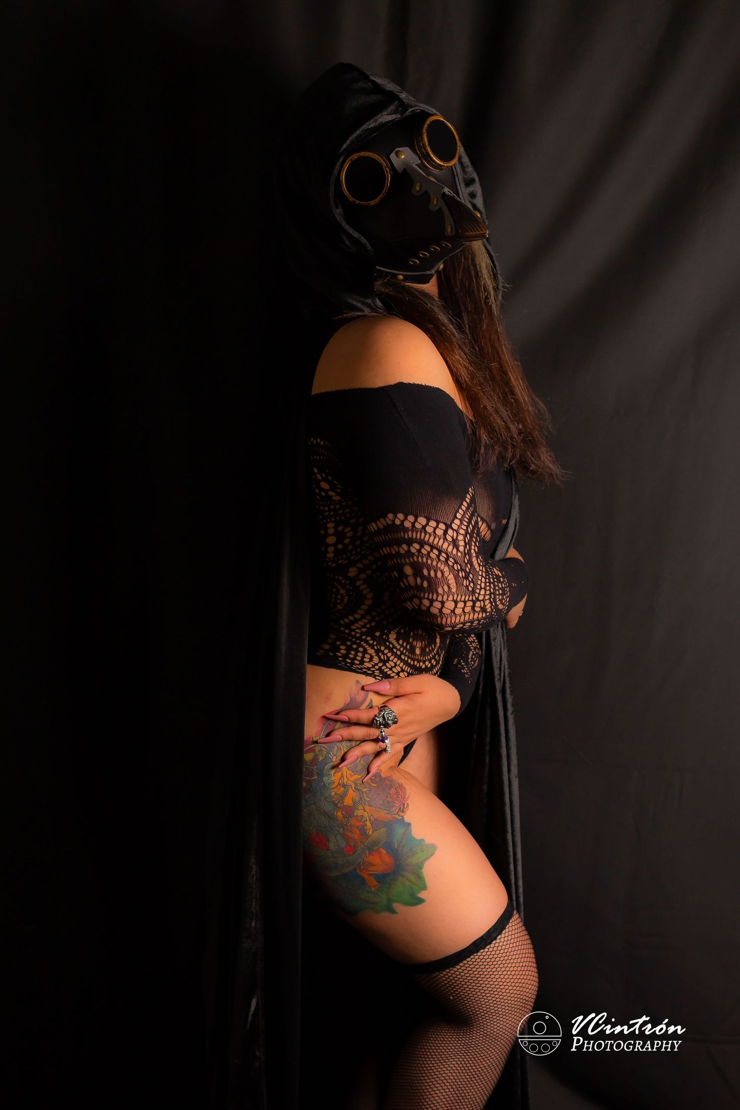 Dark artistic photo of person in black steampunk gas mask and corset with colorful thigh tattoo against dark background.