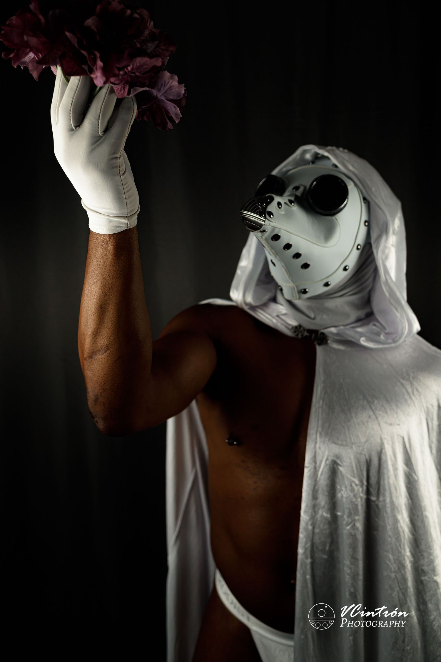 Artistic portrait showing someone in a hockey mask and white cloak holding a red flower with white gloved hand.