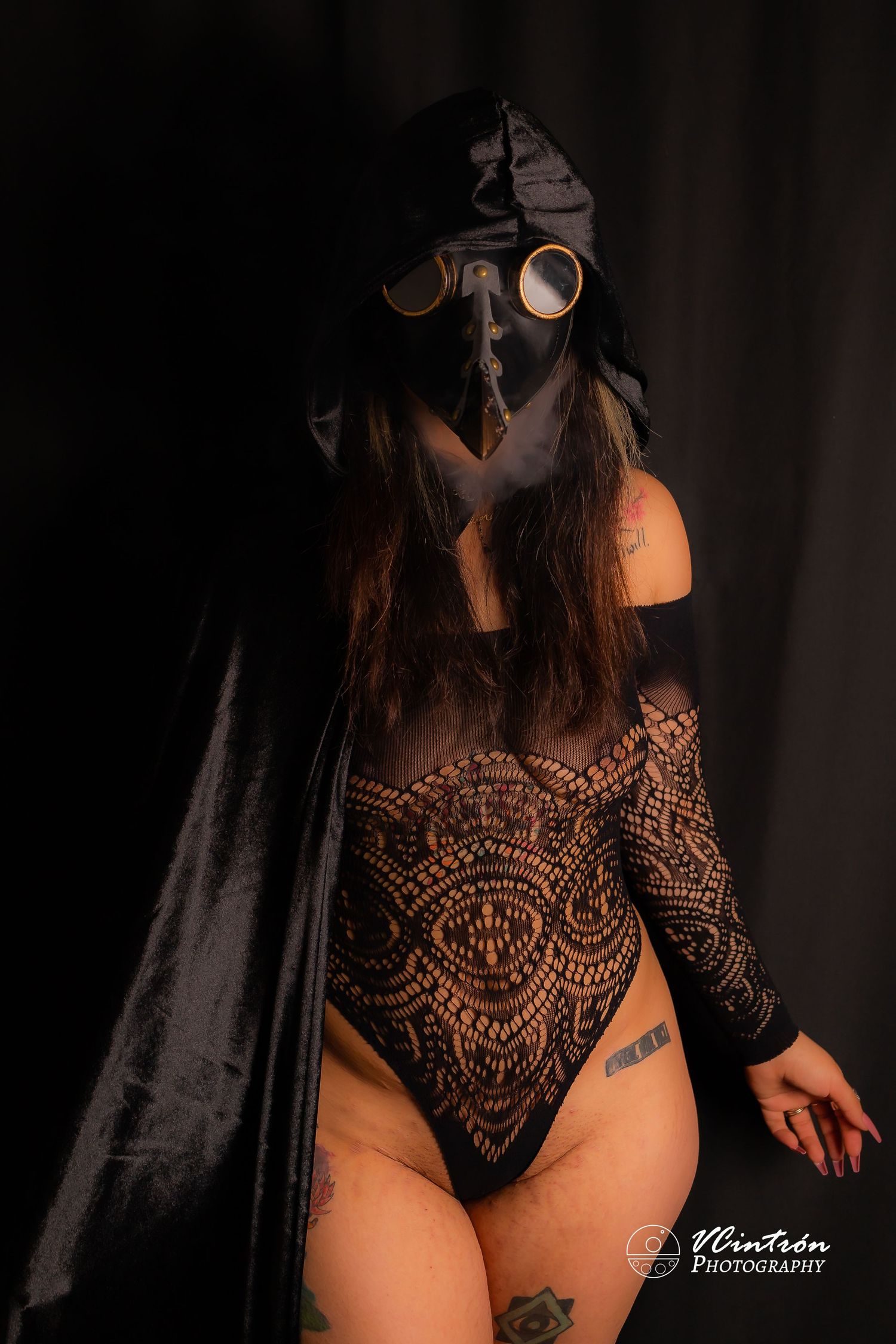 Dark artistic portrait of figure in lace bodysuit wearing gas mask and black cape in moody lighting.