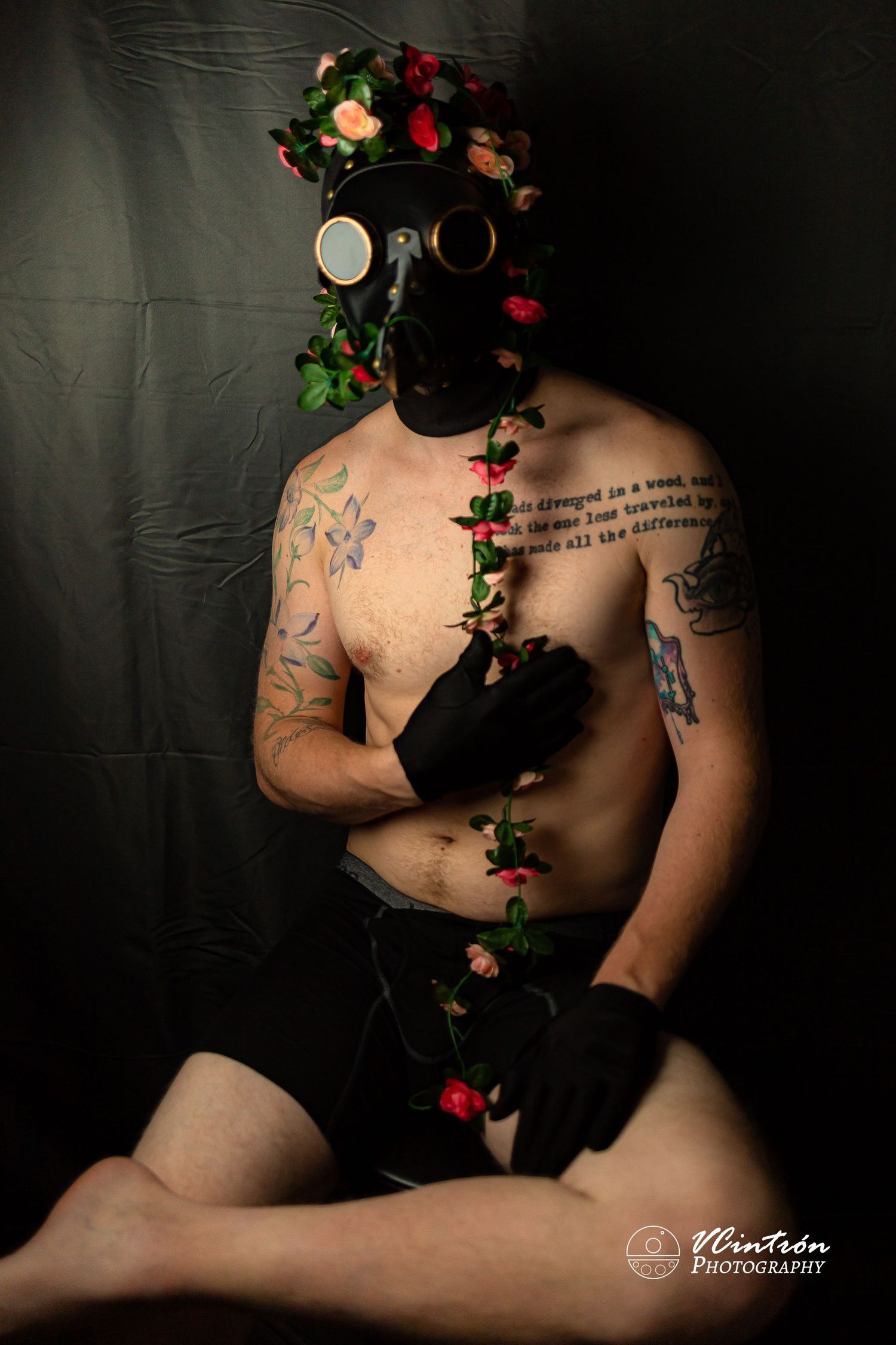 Artistic portrait featuring gas mask adorned with red roses and ivy, shirtless figure with tattoos against dark backdrop.