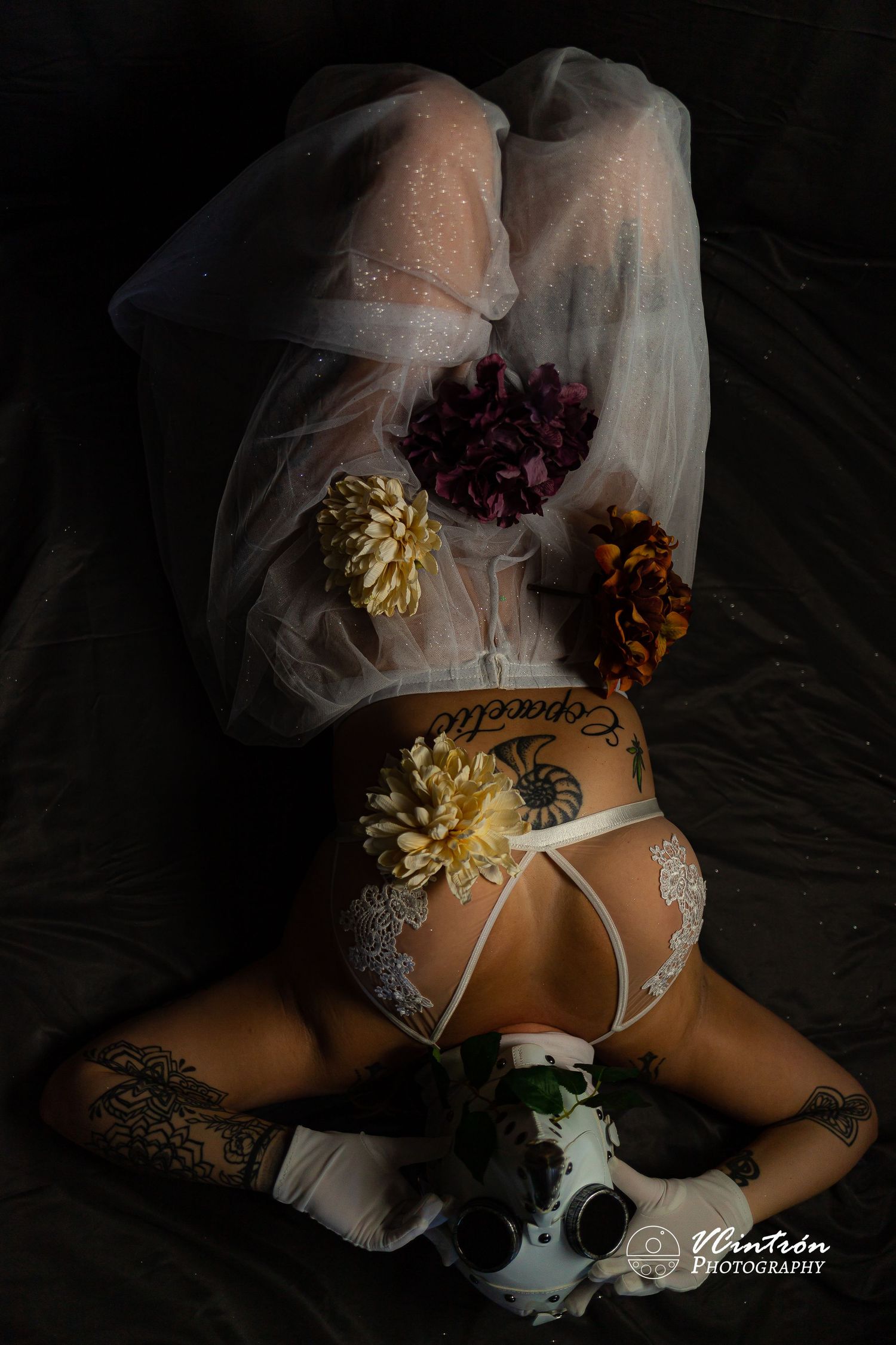 Artistic boudoir photography with white lace lingerie, veil and colorful dried flowers against dark background.