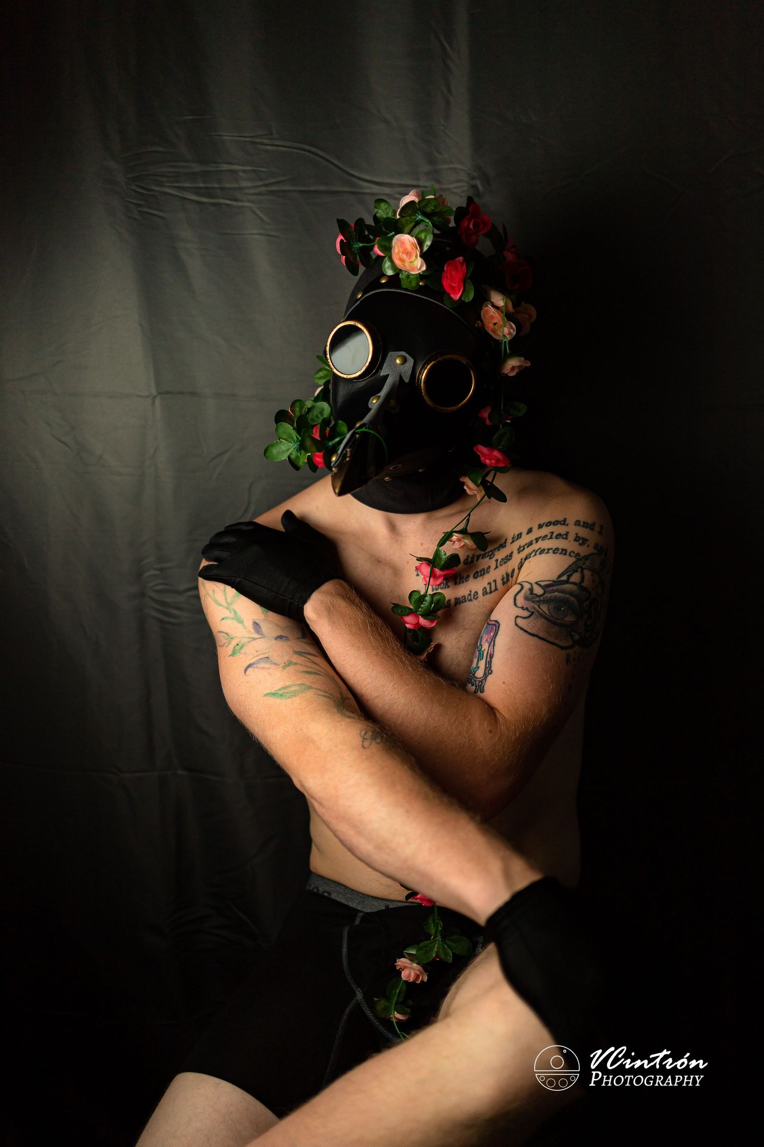 Dark artistic portrait featuring a gas mask adorned with flowers and visible arm tattoos in moody lighting.