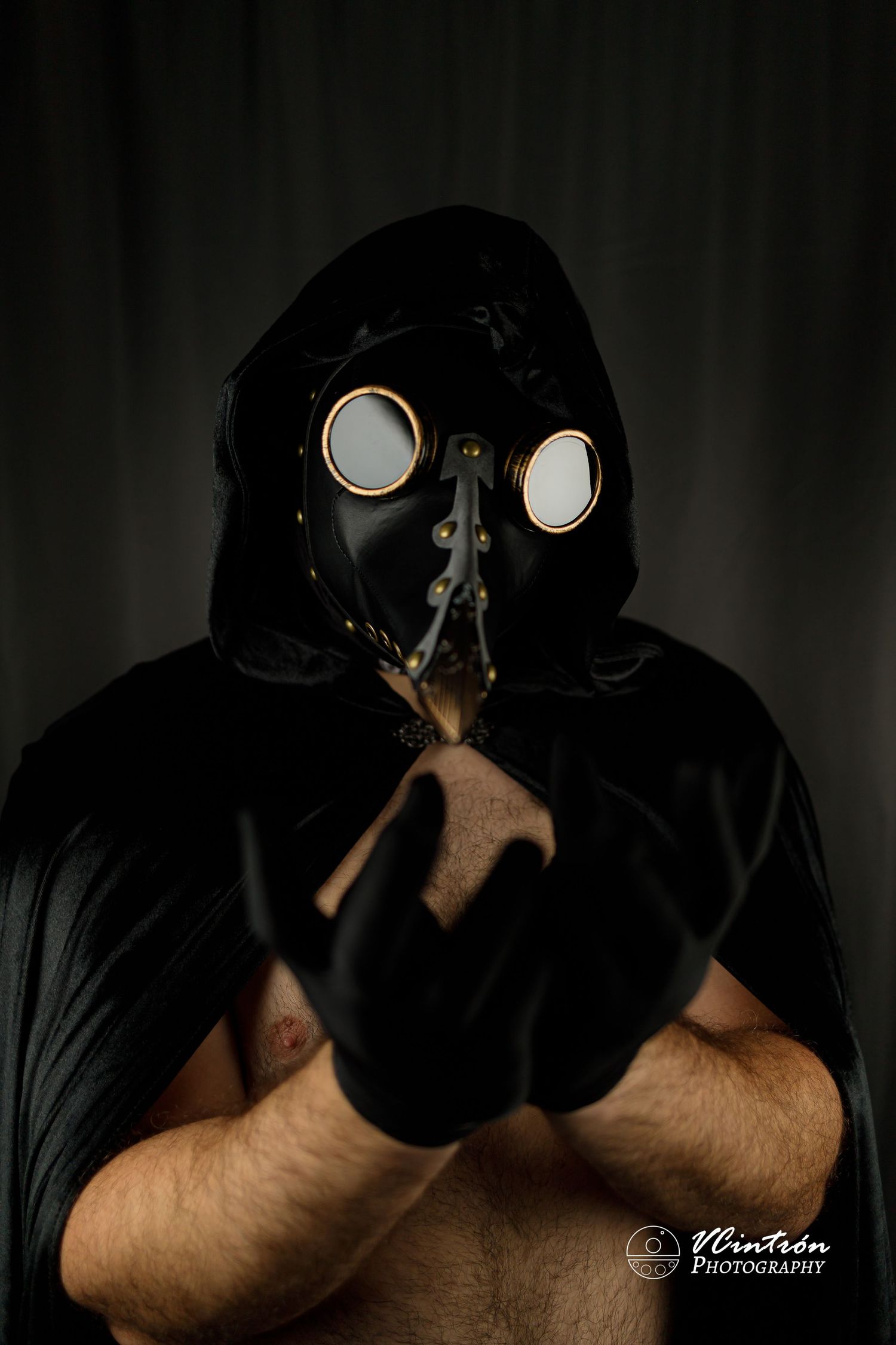 Haunting photo of a plague doctor mask figure wearing a black hooded cloak against a dark background.