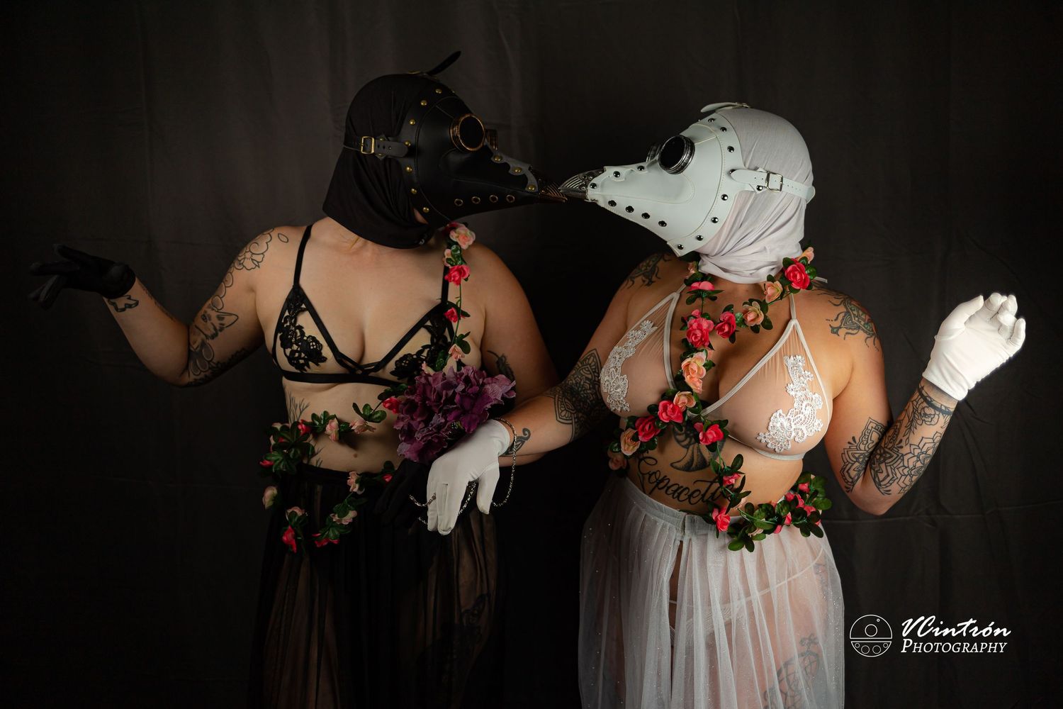 Two masked dancers in contrasting black and white lingerie costumes with floral accents perform in a dark studio setting.