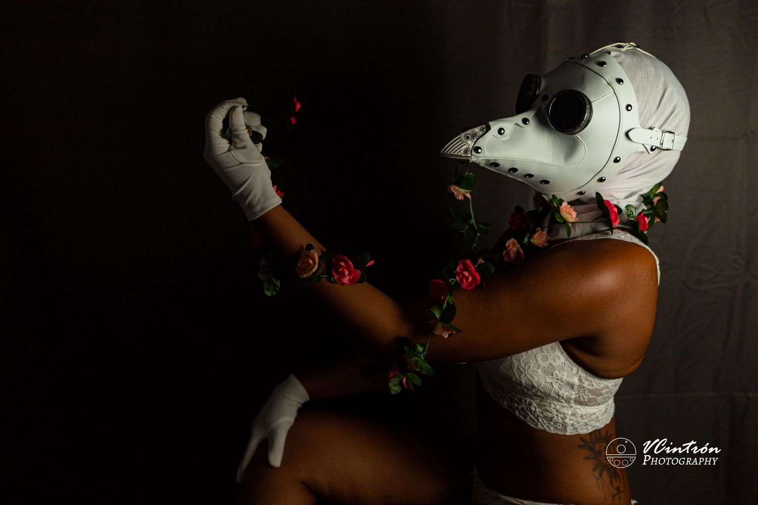 Dark artistic portrait featuring someone wearing a white plague doctor mask and floral garlands in a dramatic pose.