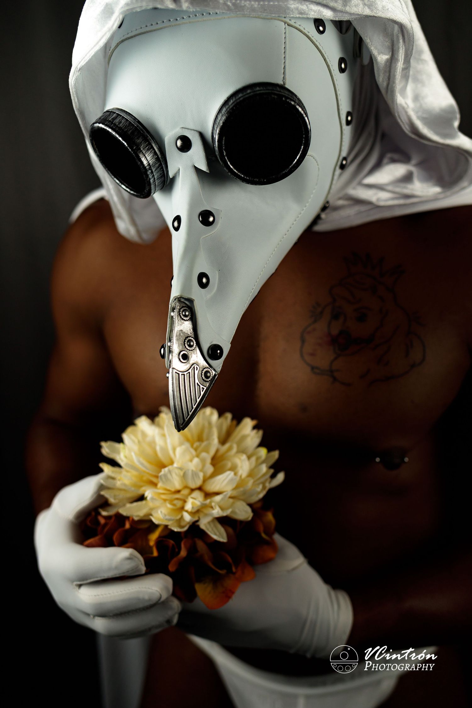 Artistic photo of someone wearing plague doctor mask and white gloves holding yellow chrysanthemum flower against dark background.