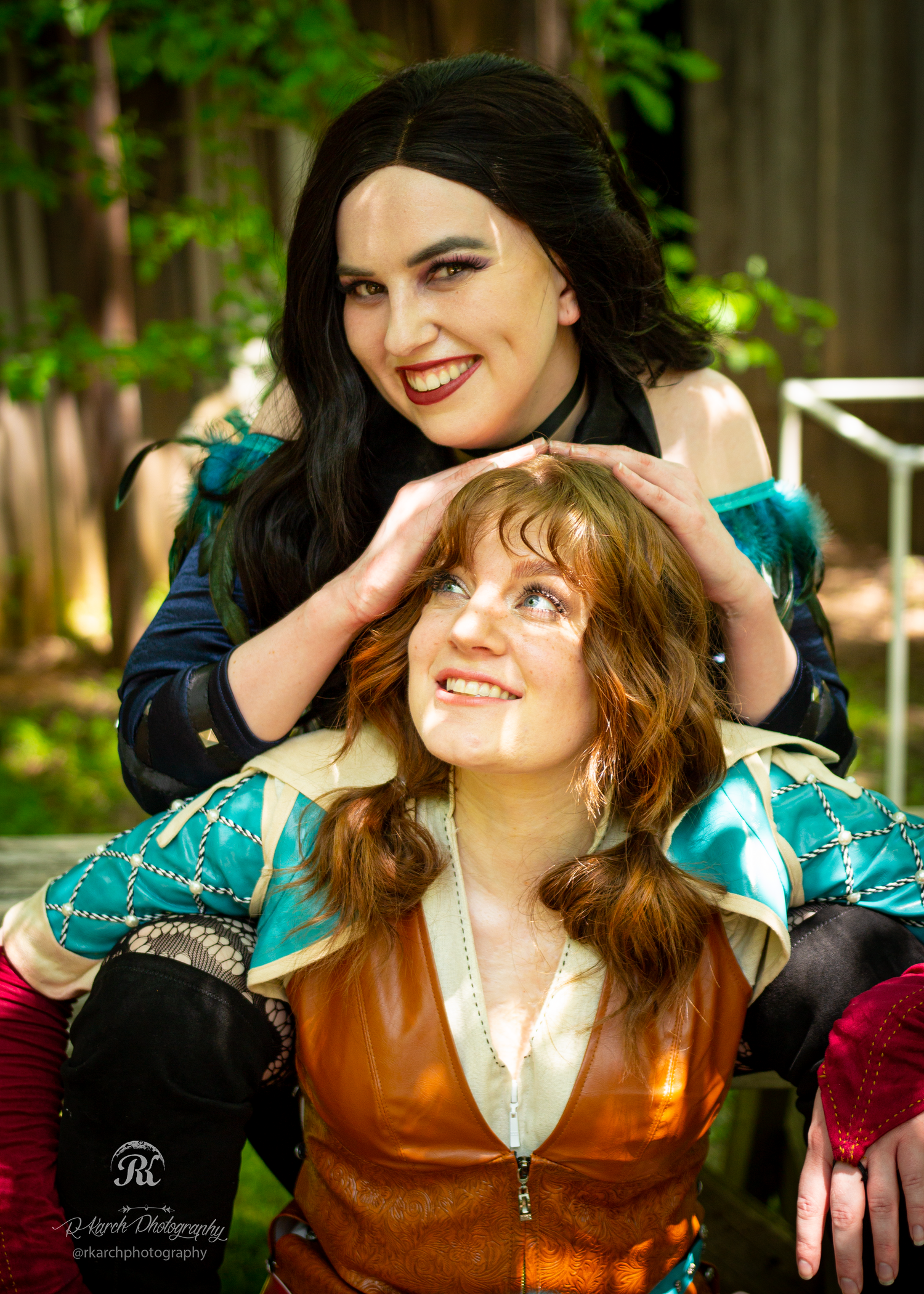 Two cosplayers in medieval fantasy costumes pose playfully in woodland setting.