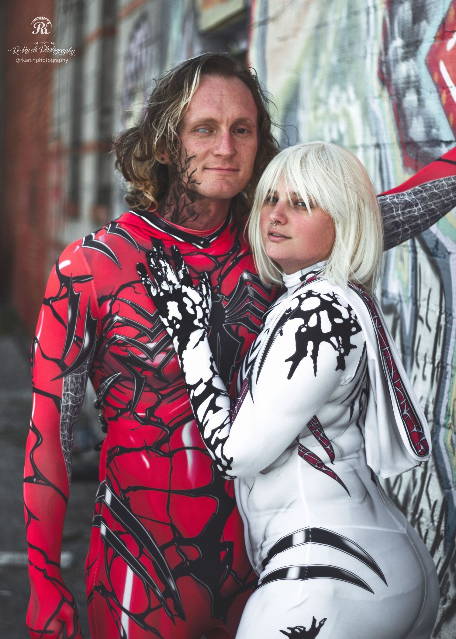 Cosplay couple in matching red and white bodysuit costumes pose against graffiti wall.