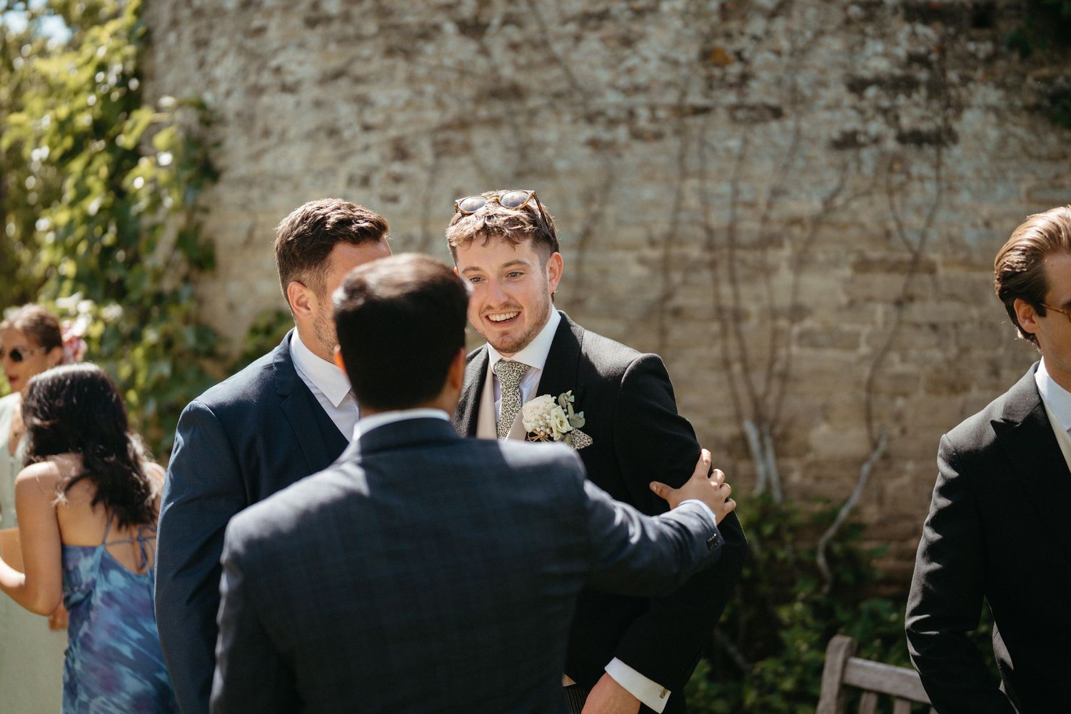 Wedding guests laugh and socialize during an outdoor celebration near a stone wall.