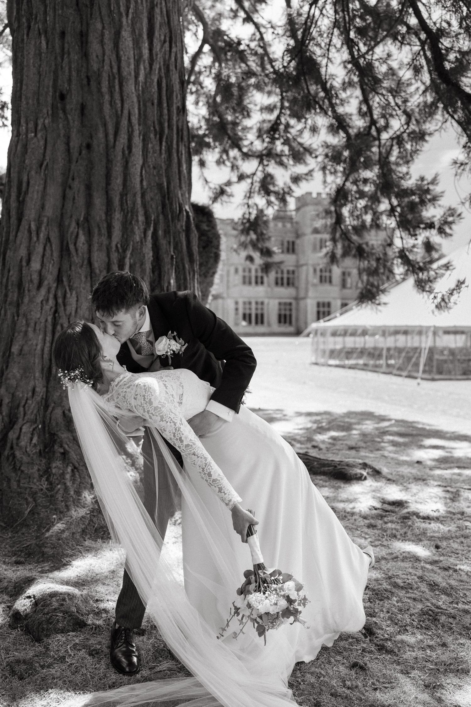 A romantic wedding photo of a couple in an elegant dip pose under a large tree with a mansion in the background.