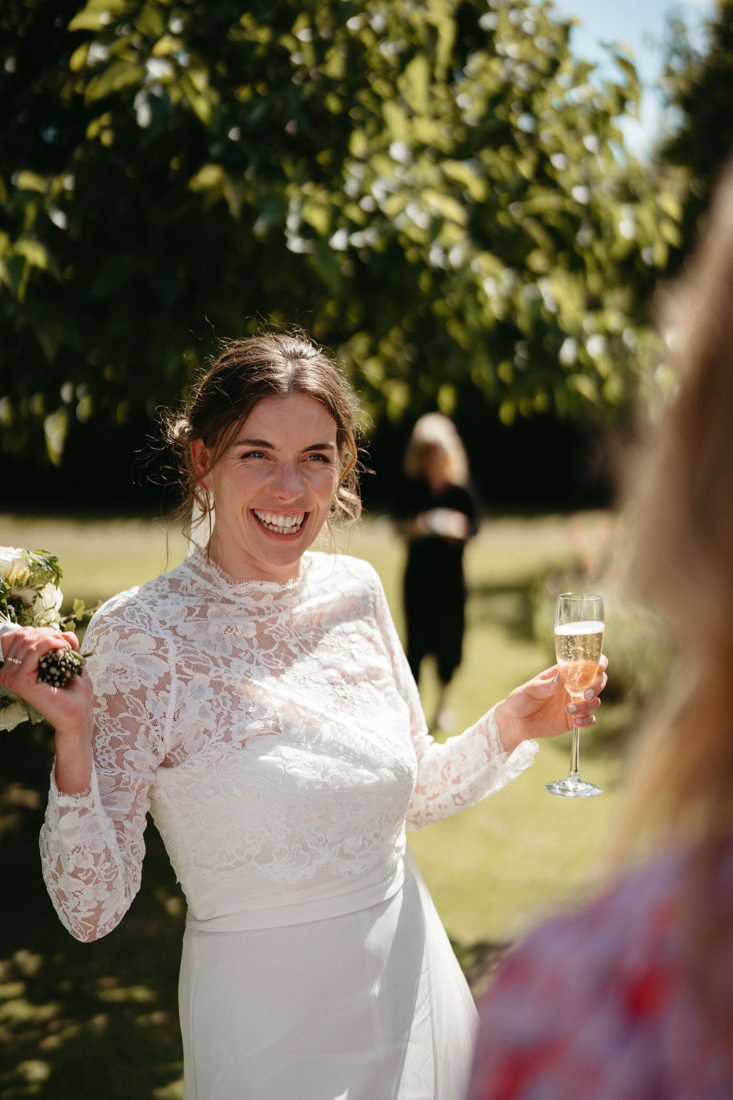 A bride in white lace raises her champagne glass while celebrating in a garden.
