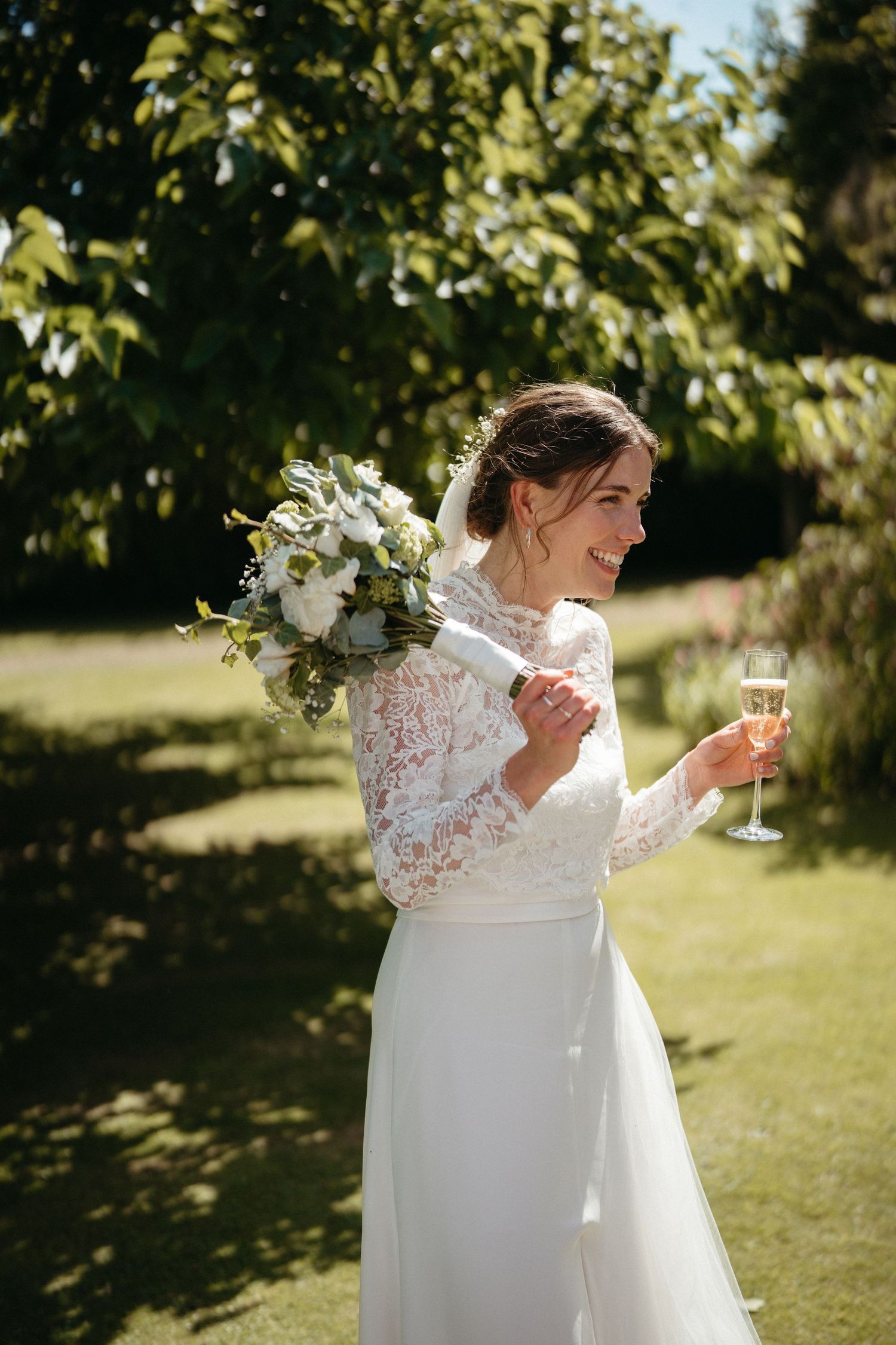 A bride in a white lace gown with long sleeves enjoys champagne in a sunny garden setting.