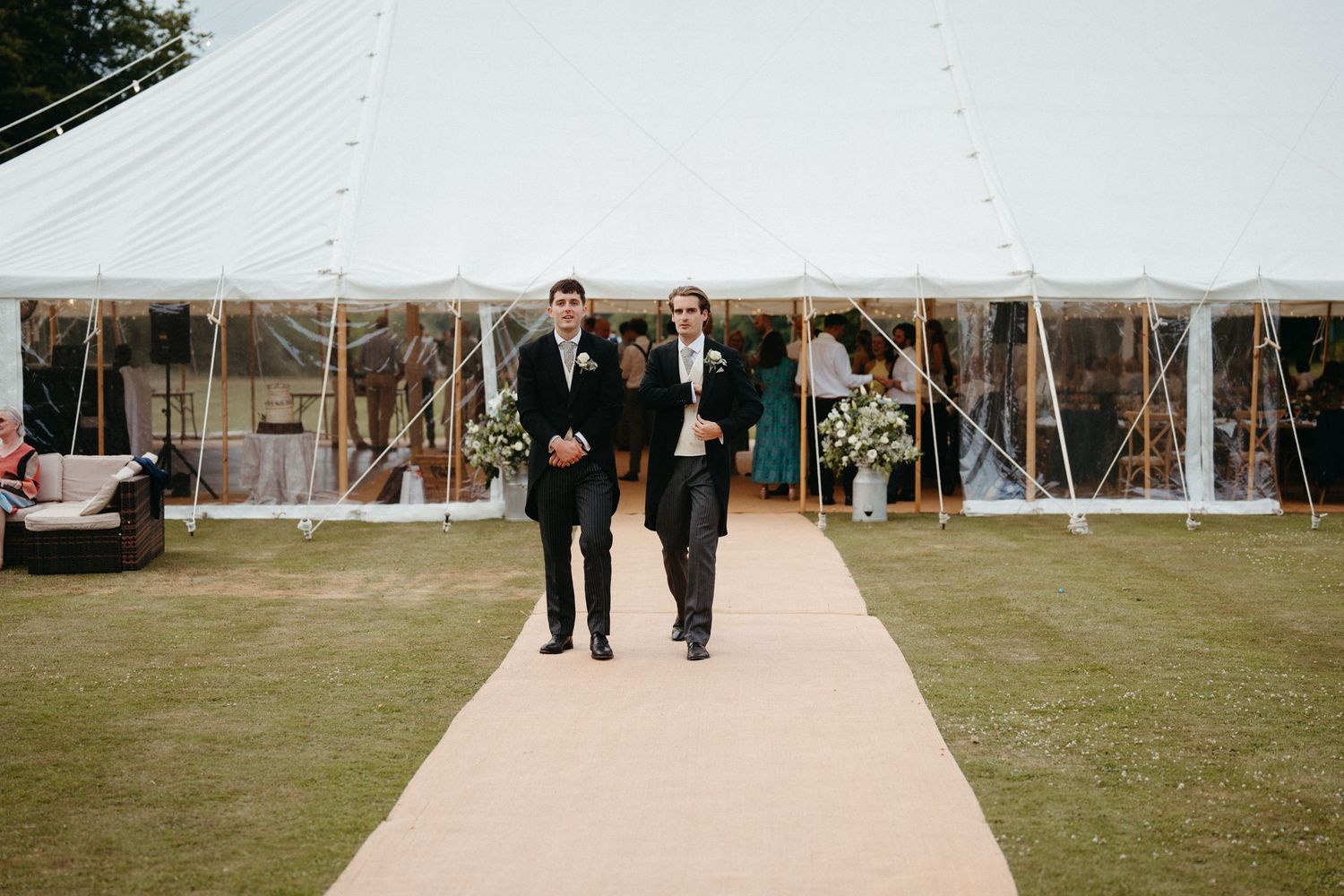 Wedding guests walking along a pathway towards a white reception tent on manicured grounds.