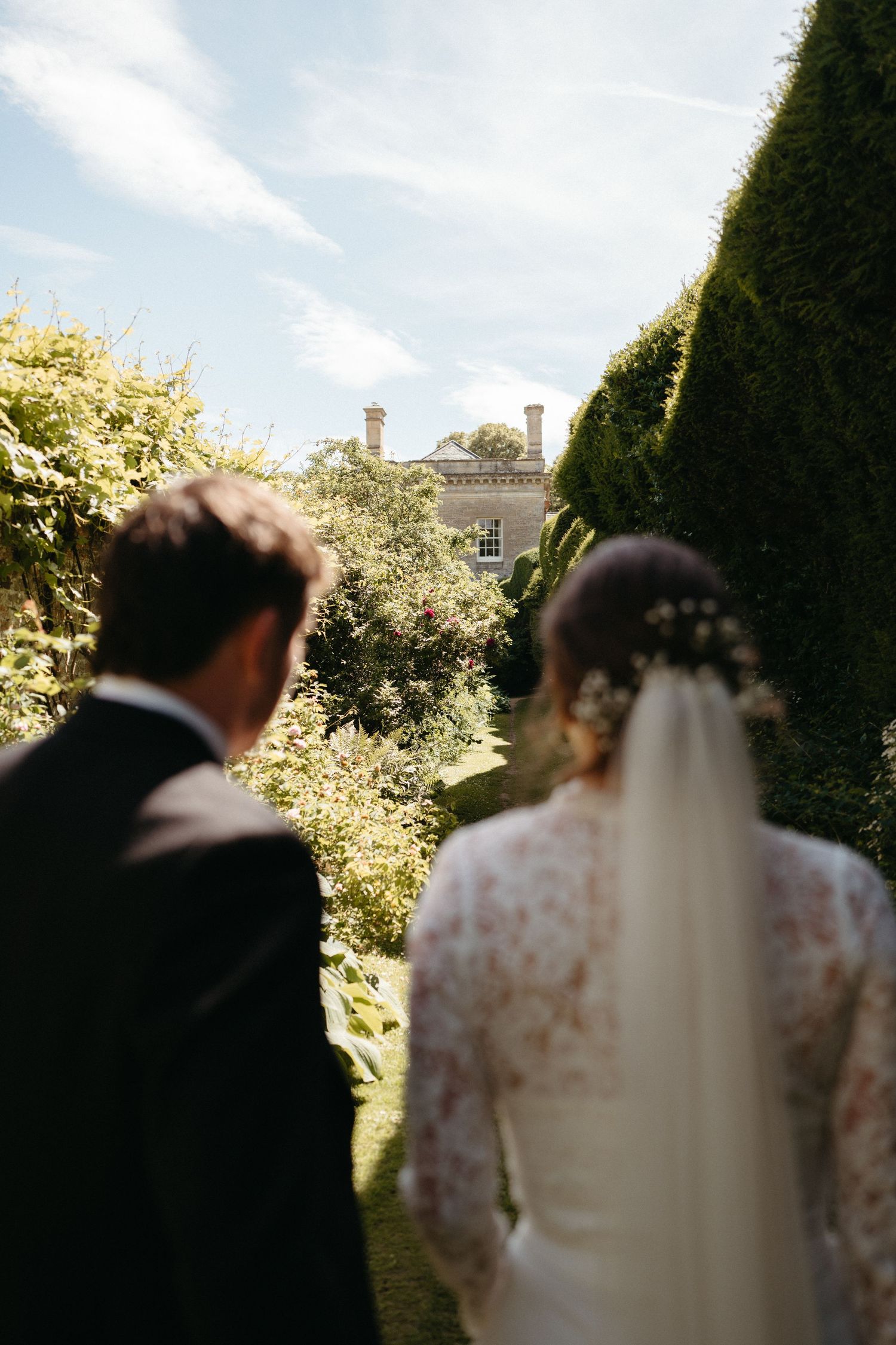 A wedding couple views a stately mansion through garden hedges on their special day.