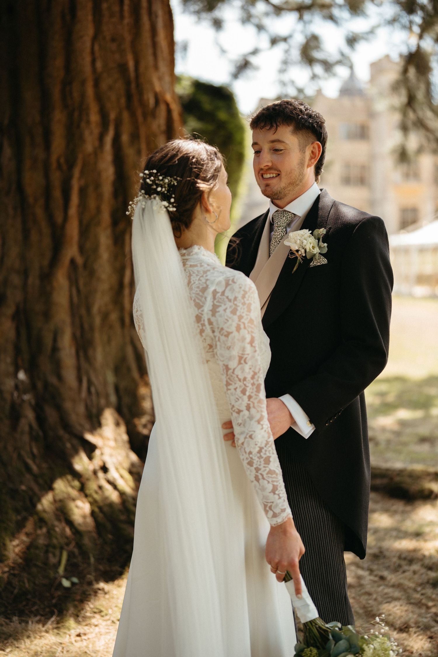 A joyful wedding couple shares laughter among towering trees in warm sunlight.