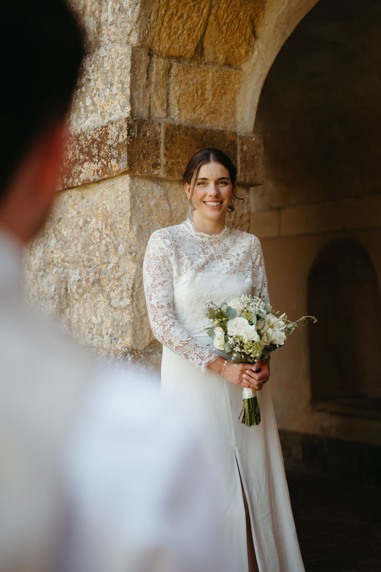 An elegant bride in white lace dress holds a bouquet of flowers in a stone corridor.