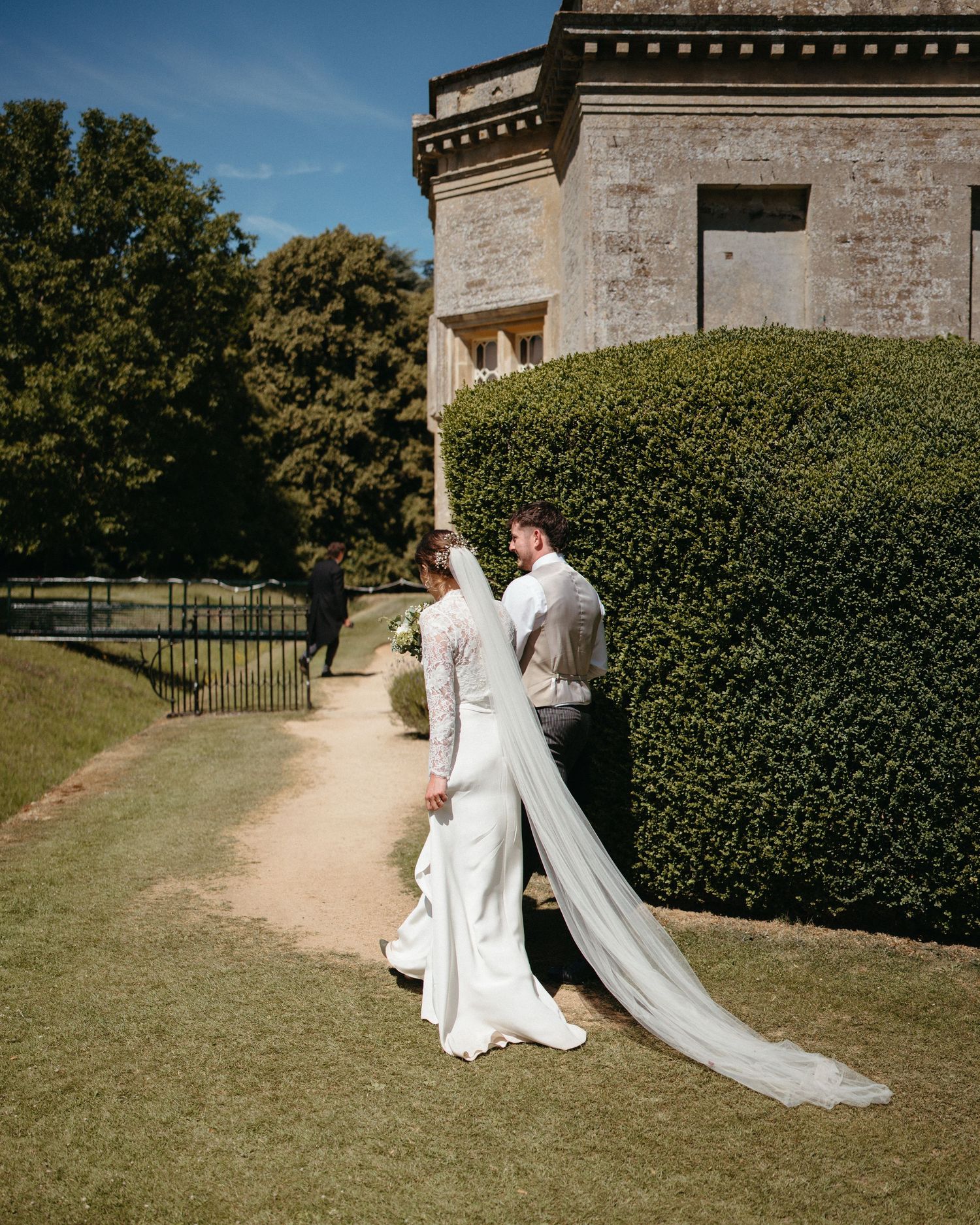 Wedding couple walks along manicured hedges at a historic stone venue.