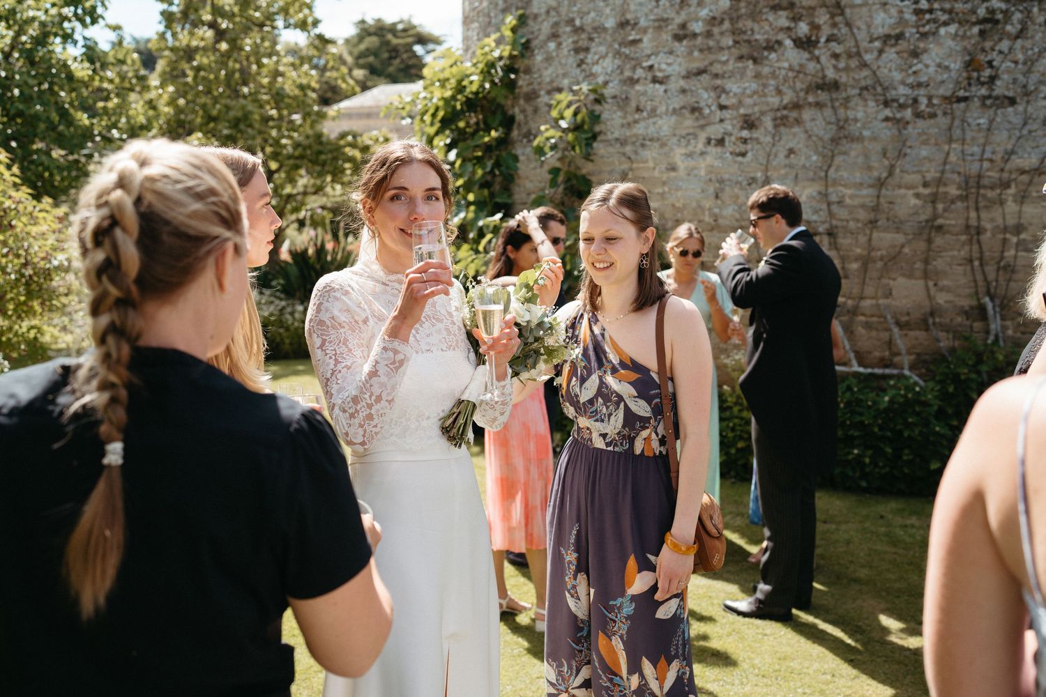 Wedding guests gather near a rustic stone wall during an outdoor reception.