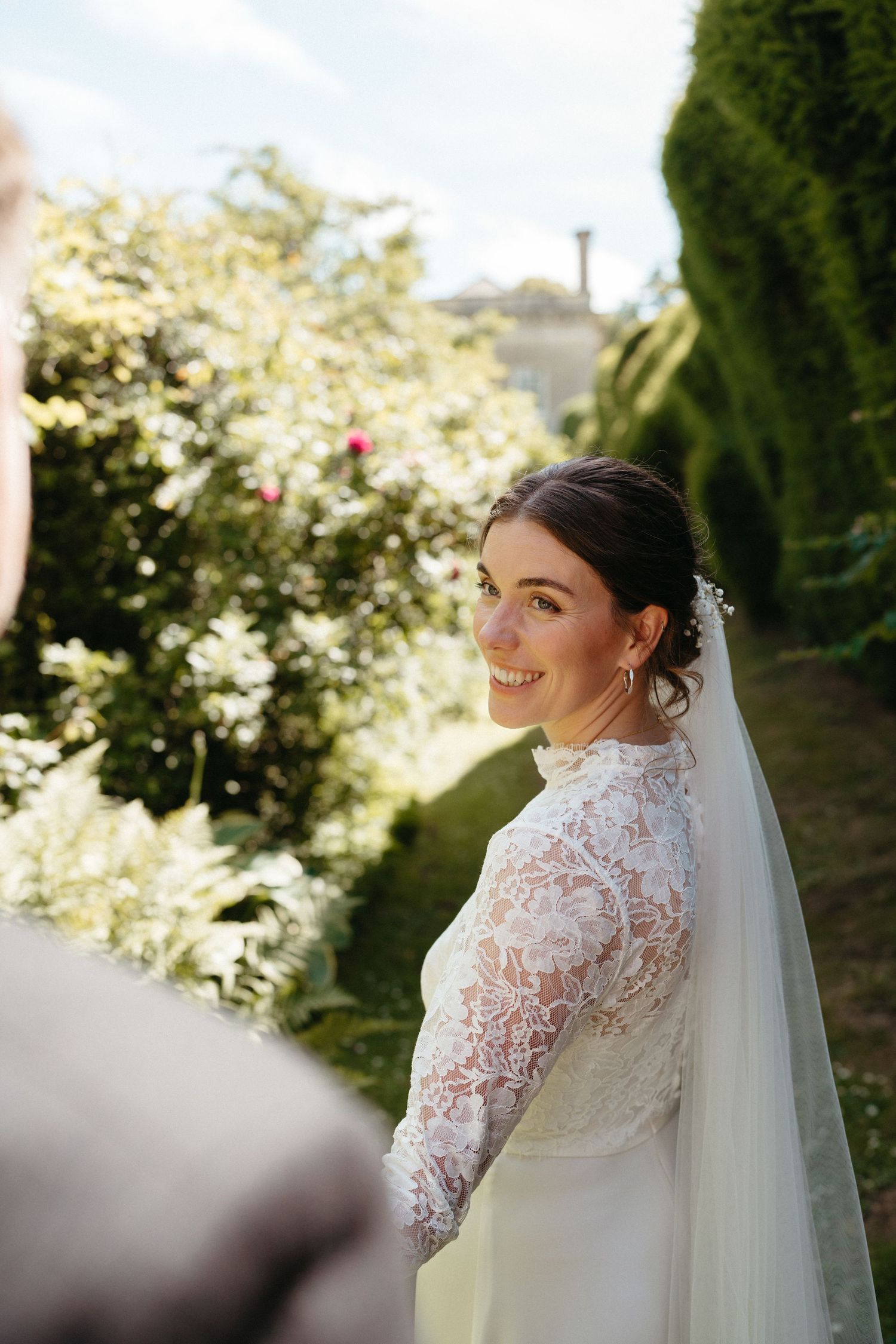 A side profile of a bride in a lace-sleeved wedding dress with flowing veil in a garden setting.