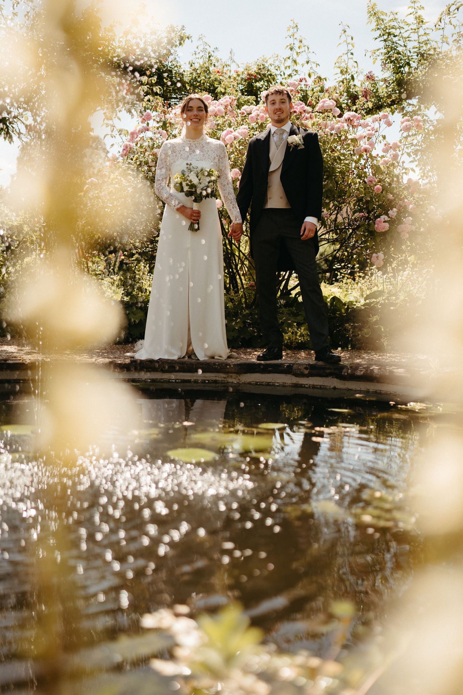 A romantic wedding portrait by a reflective pond surrounded by blooming flowers.