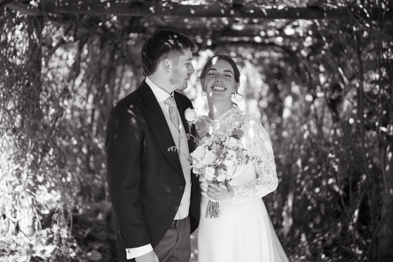 A black and white wedding portrait of a couple sharing a laugh under an ivy-covered archway.