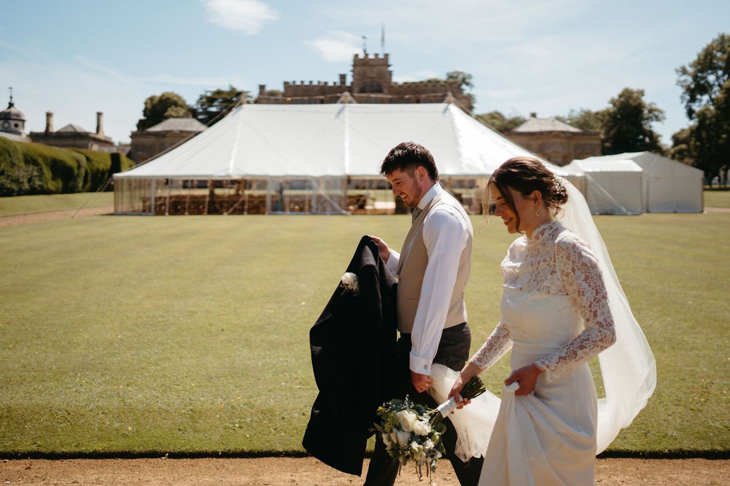 A wedding couple walks together across a sunny lawn with a large tent in the background.
