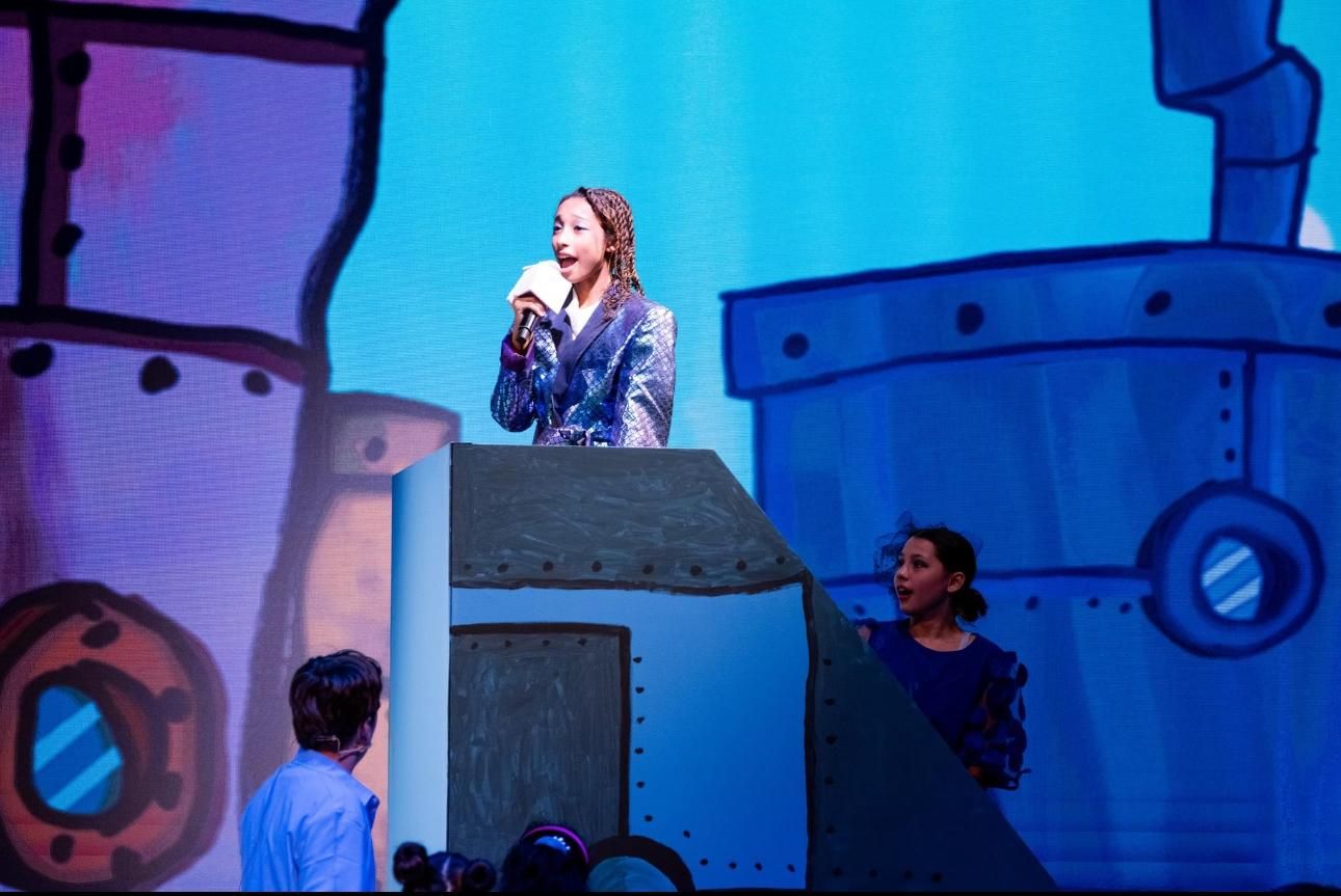 Performer on stage speaking into microphone against vibrant blue backdrop with theatrical lighting effects.