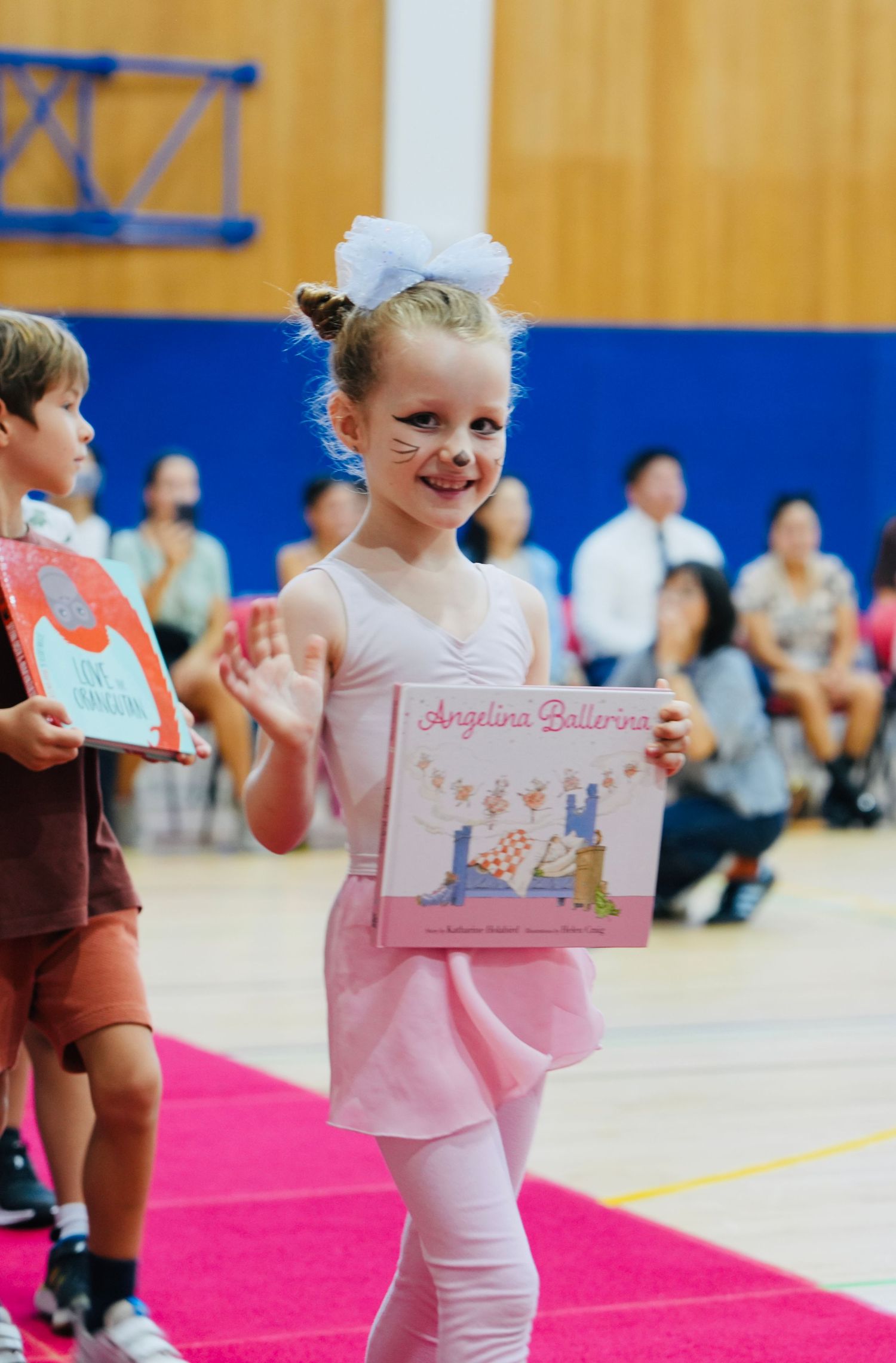 Young dancers in pink tutus perform on a bright pink mat in a school gymnasium.