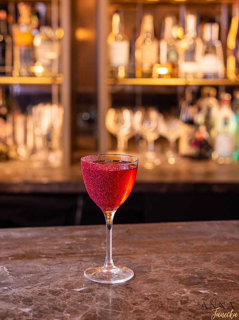 Vibrant red cocktail served in a wine glass against blurred bar shelving background.