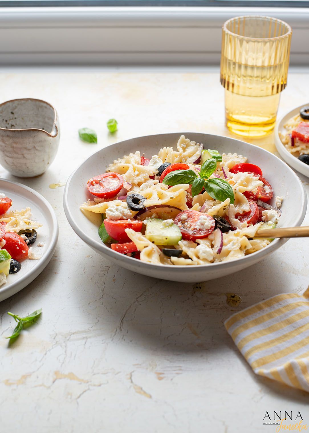 Fresh pasta salad with tomatoes, olives and herbs served in a white bowl with yellow glassware.