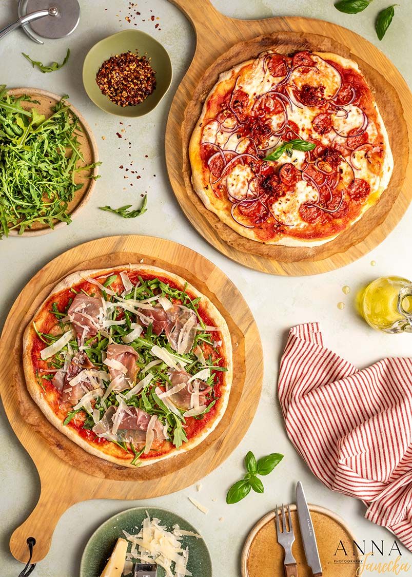 Two gourmet pizzas topped with prosciutto, arugula and fresh mozzarella on wooden serving boards.