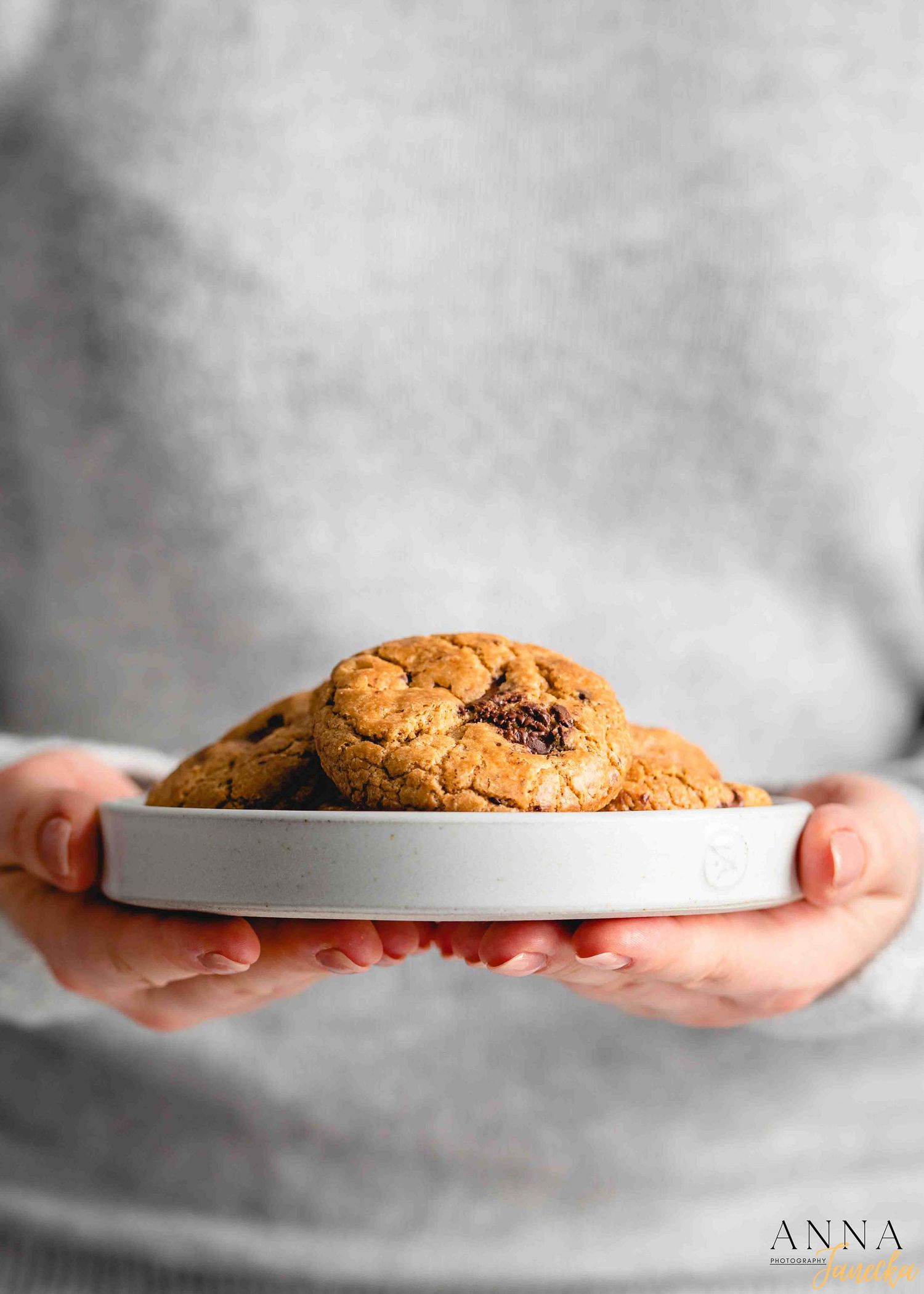 Hands holding a large chocolate chip cookie on a white plate against a gray background.