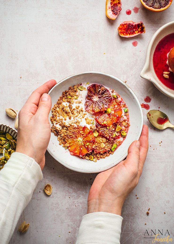 Hands holding a bowl of colorful breakfast porridge topped with blood oranges, nuts and seeds.