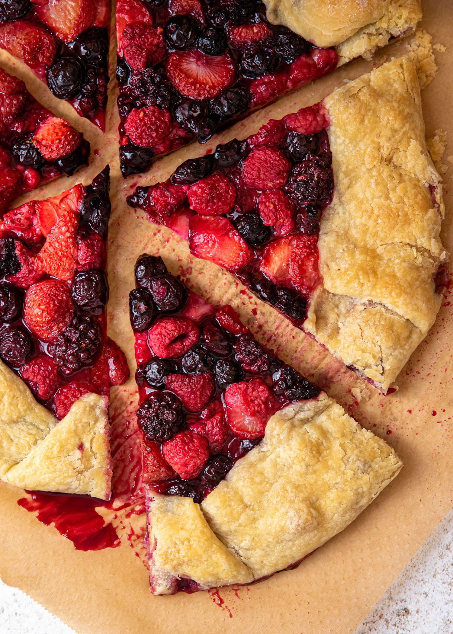 A rustic mixed berry galette with golden flaky crust filled with fresh raspberries and blueberries on parchment paper.