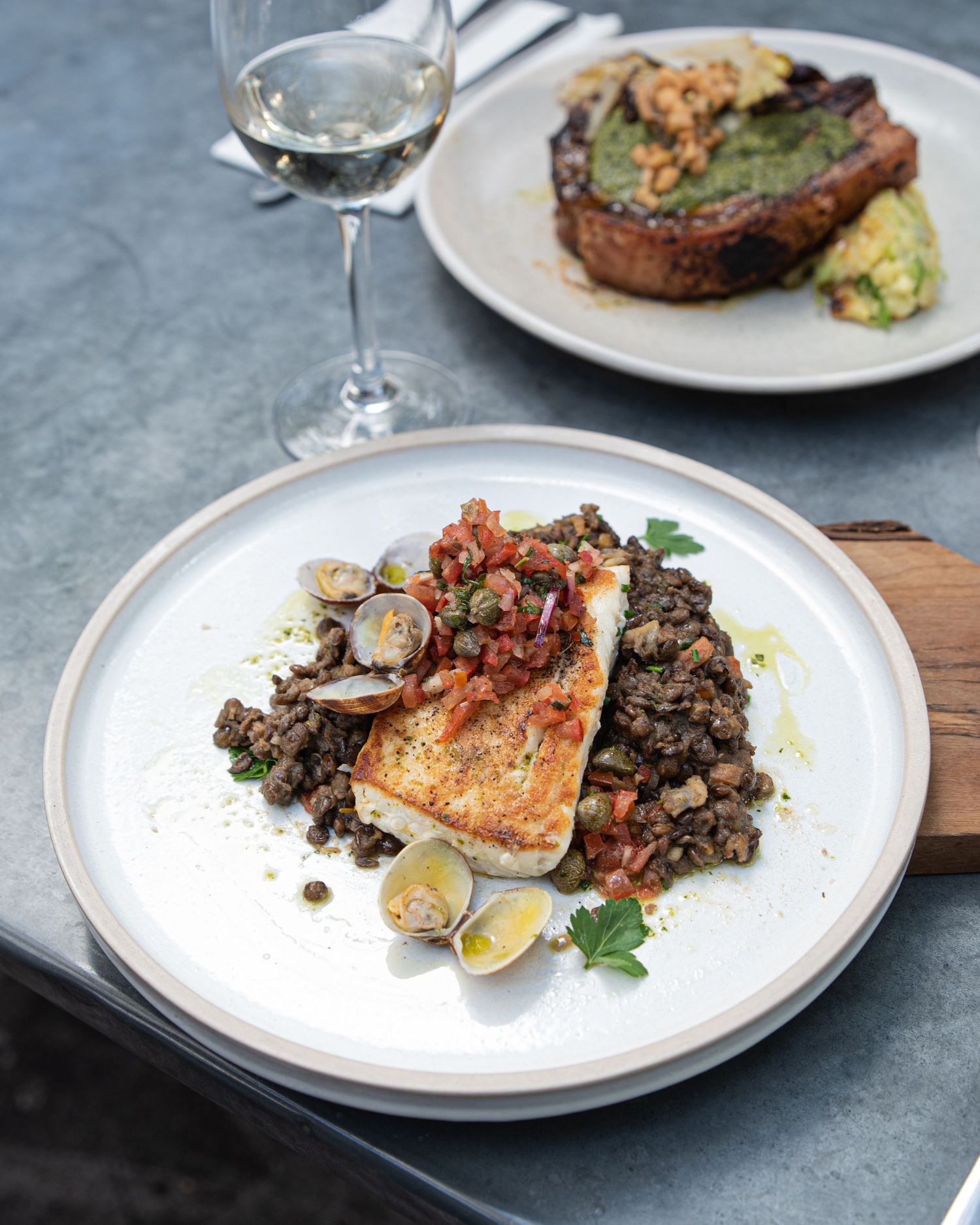 Pan-seared fish served with lentils and clams on white plates alongside a glass of white wine.