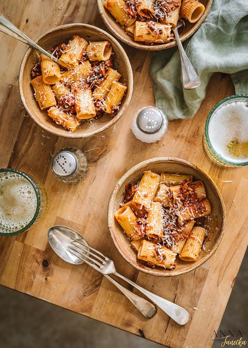 Three bowls of rigatoni pasta with red sauce and grated cheese served alongside cold beers on a wooden table.