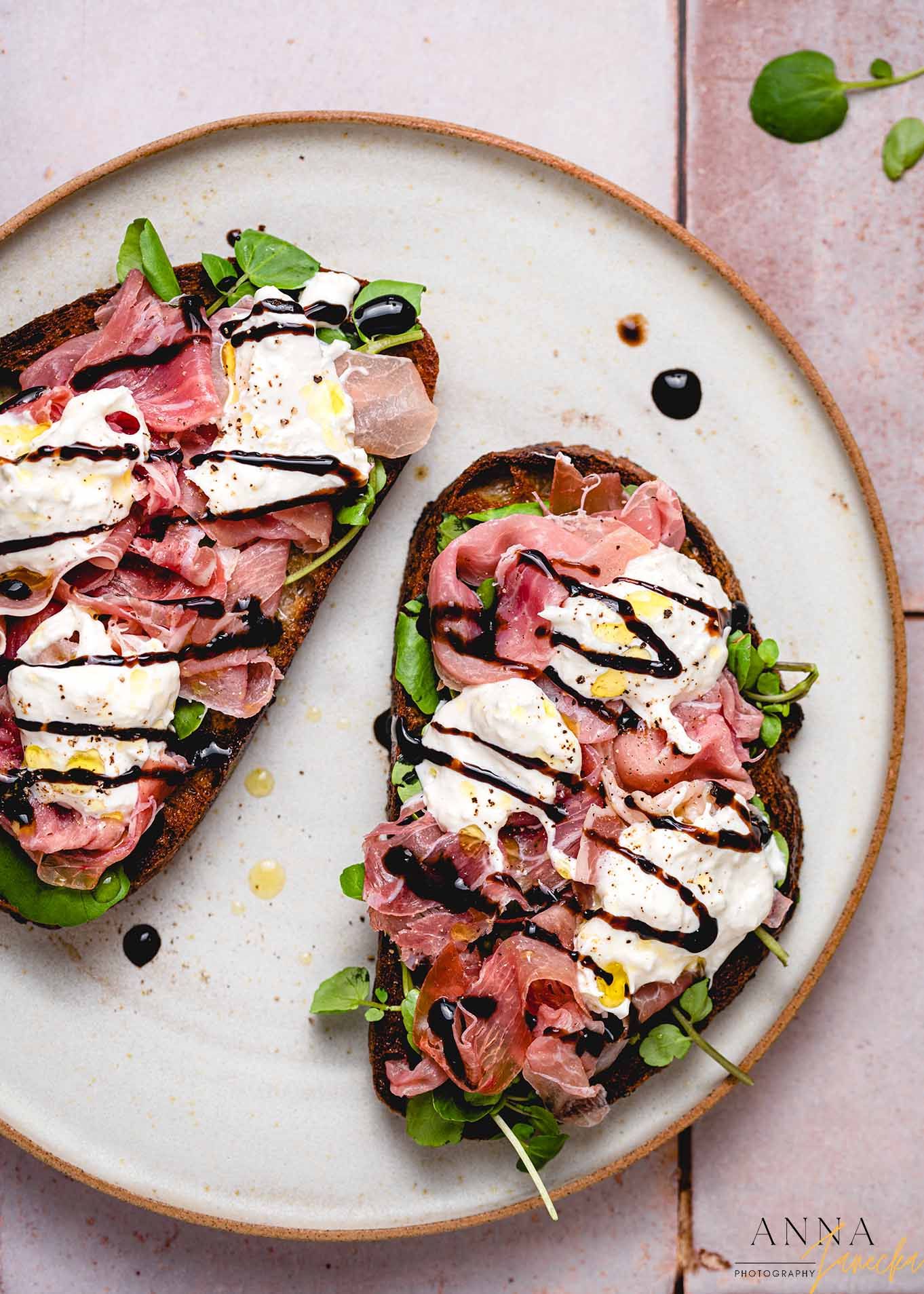 Gourmet open-faced sandwich with prosciutto, burrata, and balsamic drizzle on white plate.