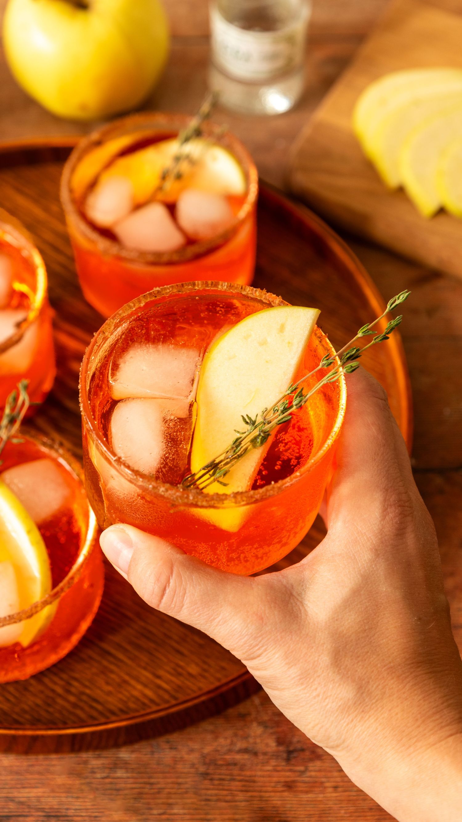 Orange cocktail garnished with lemon wedge and thyme sprig served on wooden tray with ice.