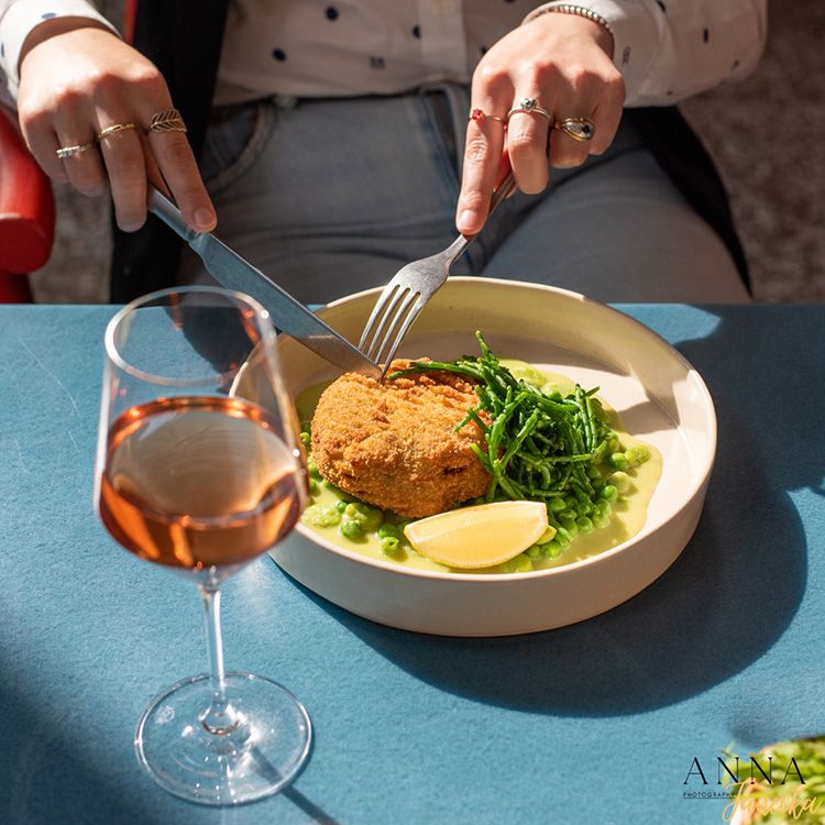 Elegant plated crab cake with fresh greens and glass of rose wine on blue tablecloth.