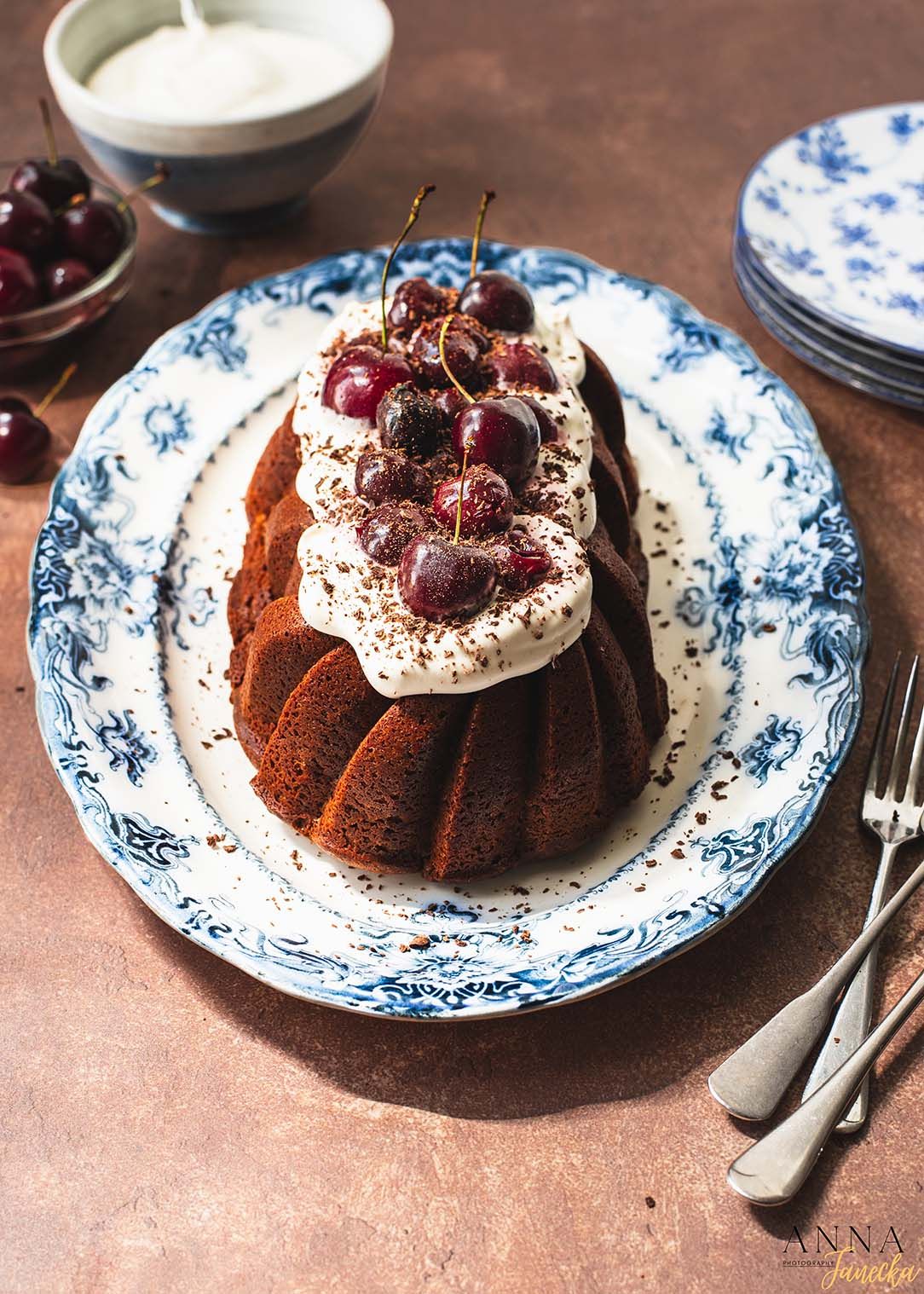 Chocolate bundt cake topped with whipped cream and fresh cherries on blue and white porcelain plate.