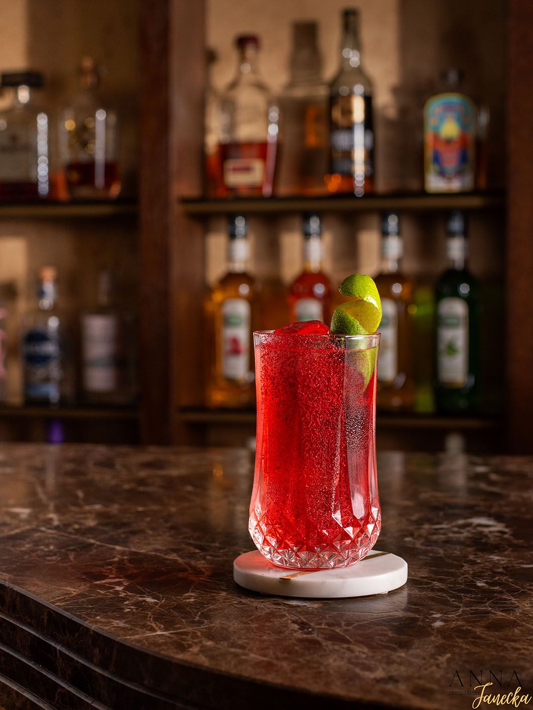 Red sparkling cocktail with lime garnish served on marble bar counter with bottle display behind.